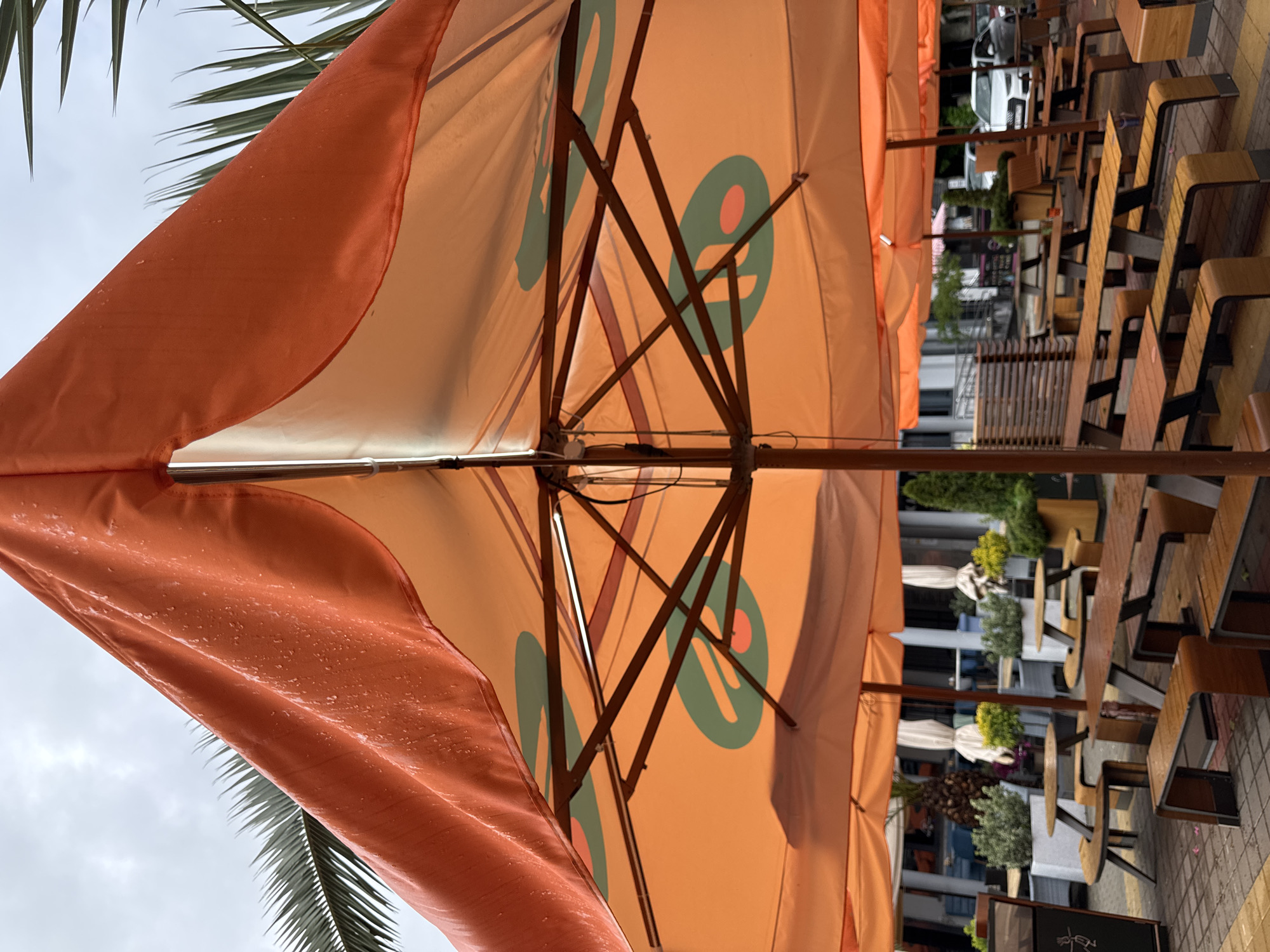 This image depicts an outdoor seating area, likely a cafe or restaurant, in Sochi, Russia. The perspective is from underneath a row of orange umbrellas, each bearing a circular logo with a distinctive design. Raindrops are visible on the umbrella closest to the camera, indicating recent or ongoing precipitation. The seating consists of wooden tables and benches/chairs, arranged neatly along the pavement. In the background, blurred details of the city are visible, including buildings, greenery, and parked cars. There are no people clearly visible in the image.