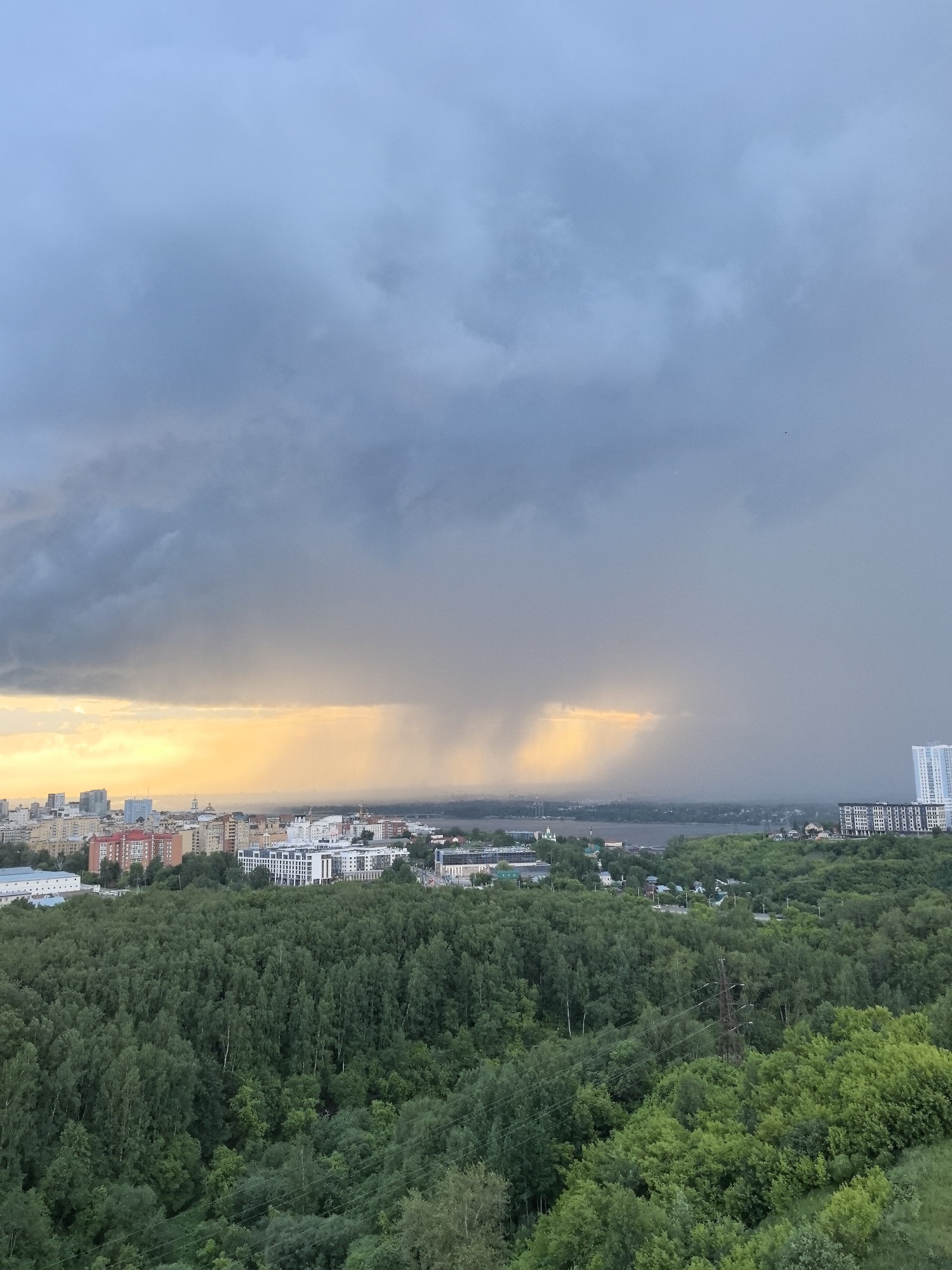 An aerial view captures a city skyline at sunset, with a dramatic storm approaching from the distance. The foreground is dominated by a dense forest, its canopy a vibrant green. Beyond the trees, a cityscape unfolds, featuring various buildings, some with colorful facades, and a distinct tall, modern residential building. A large body of water, likely the Kama River, is visible in the mid-ground, separating the city from a distant forested area. The sky is a mix of dark, ominous storm clouds and a bright, golden sunset glow where the sun's rays are breaking through. A visible rain shaft descends from the clouds, creating a curtain of water in the distance, obscuring the horizon. The overall scene suggests a transition from fair weather to a storm. The location is identified as Perm, Russia. No people are visible in the image.