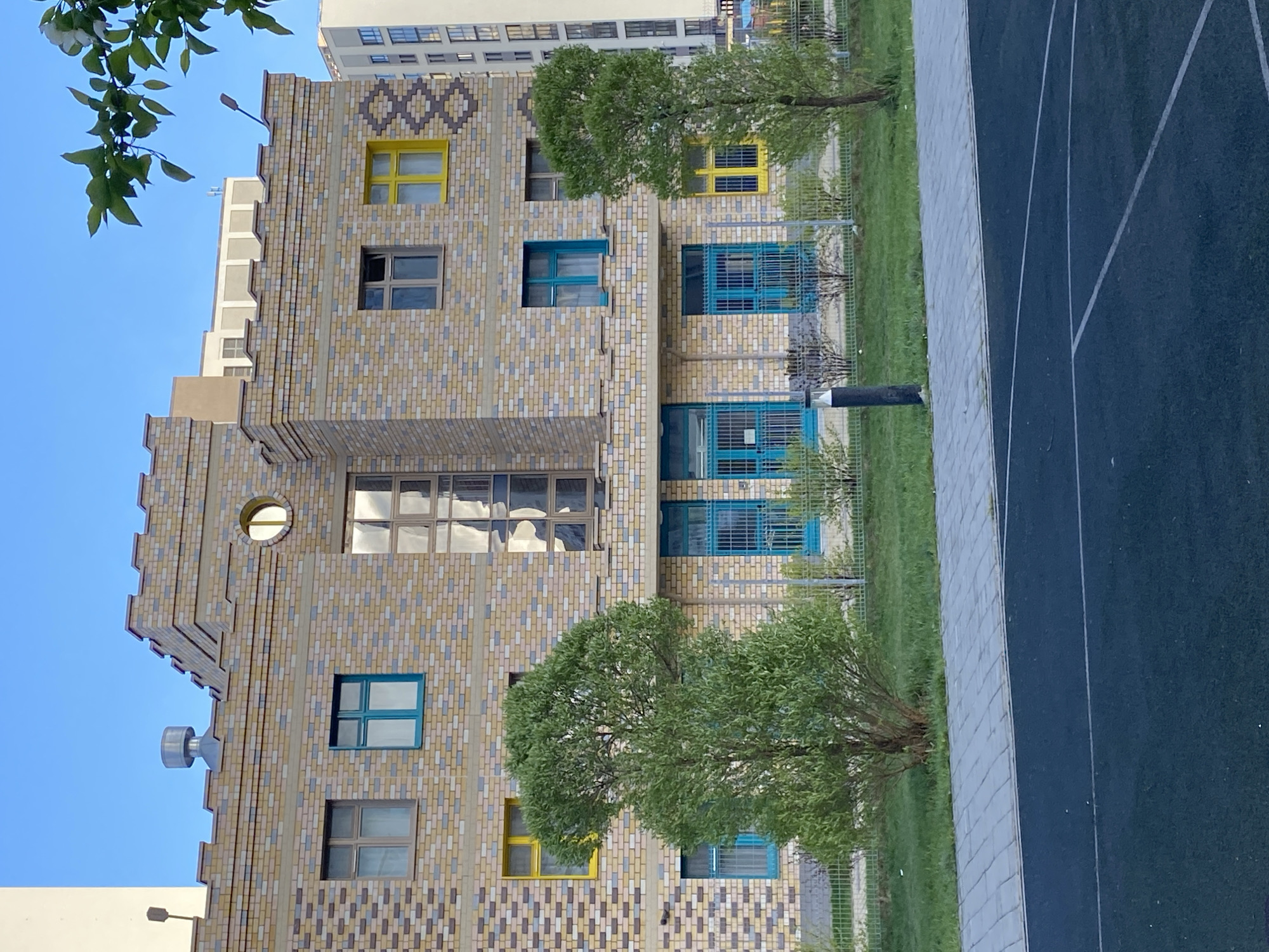 The image shows a building in Yekaterinburg, Russia, with a brick facade, windows with blue and yellow frames, and trees in front. The building has a distinctive stepped design on its left side, with a circular window and a metal vent. To the right of the building is a green space and a paved area with painted lines, possibly a sports field or track. The sky is clear and blue. Another building is visible in the background.