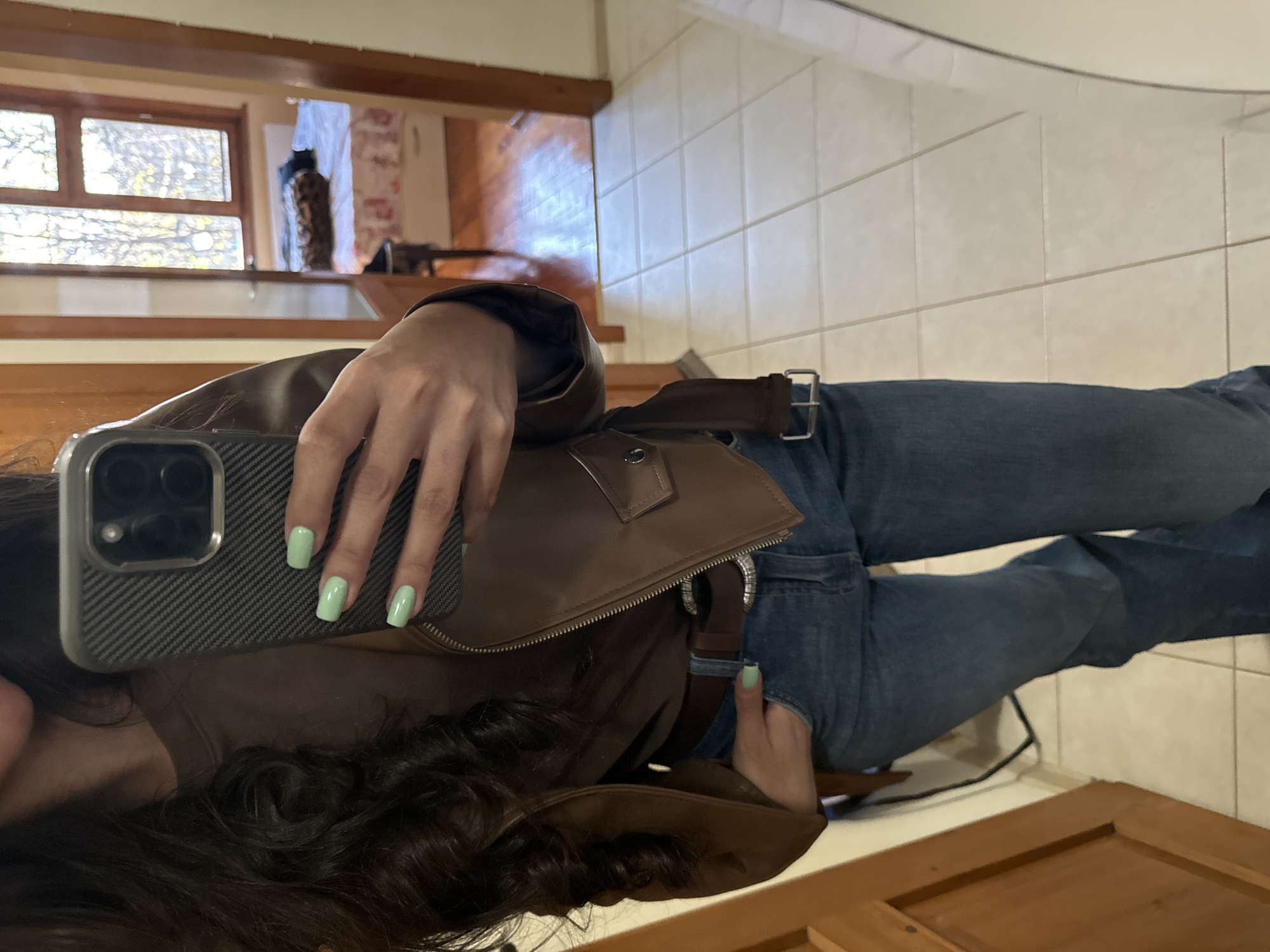 A person with dark curly hair is taking a selfie with an iPhone. The person is wearing a brown leather jacket, blue jeans with a brown belt. The person has light green nail polish. The background includes a staircase, a window with a view of trees, and tiled walls. The location cannot be determined to be Dupnitsa, Bulgaria, based on the visual information provided.