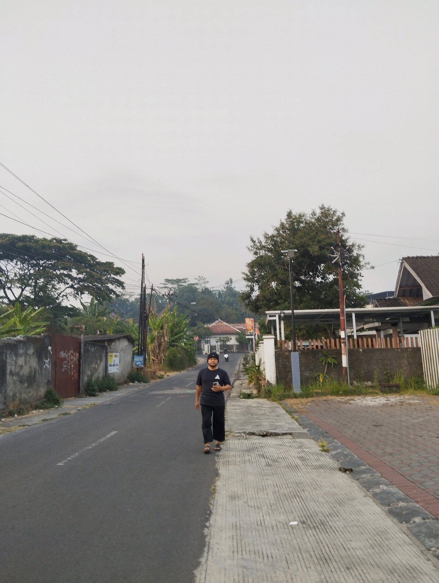In the city of Salatiga, Indonesia, a man walks on the right side of a road, which is paved and lined with a sidewalk. He is wearing a black t-shirt and black pants, and he is holding a dark-colored object in his right hand. The man is walking away from the viewer, towards a slight incline in the road. To the left of the road, there is a concrete wall with graffiti, a rusty gate, and a small structure. Behind the wall, there are trees and houses. To the right of the road, there are houses, fences, and more trees. The sky is overcast.