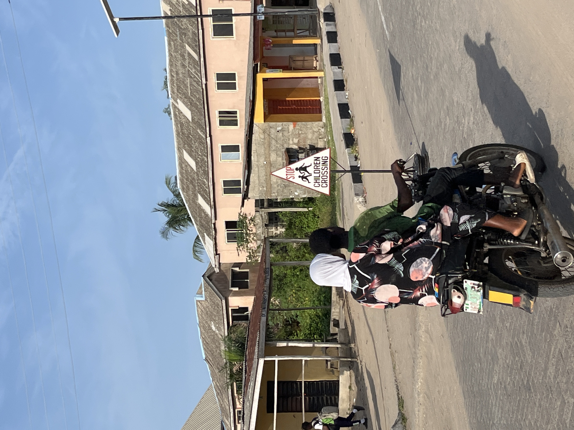 The scene is a street in Badagry, Nigeria, on a sunny day. A man is riding a motorcycle on the right side of the road. A passenger in a black shirt with white patterns is seated behind him. The motorcycle has a license plate and a small red item attached to the back. A triangular "Stop Children Crossing" sign is visible near a building. The buildings are light pink, yellow, and grey. The sky is clear and blue with utility poles and wires. The shadow of the motorcycle rider is visible on the street.