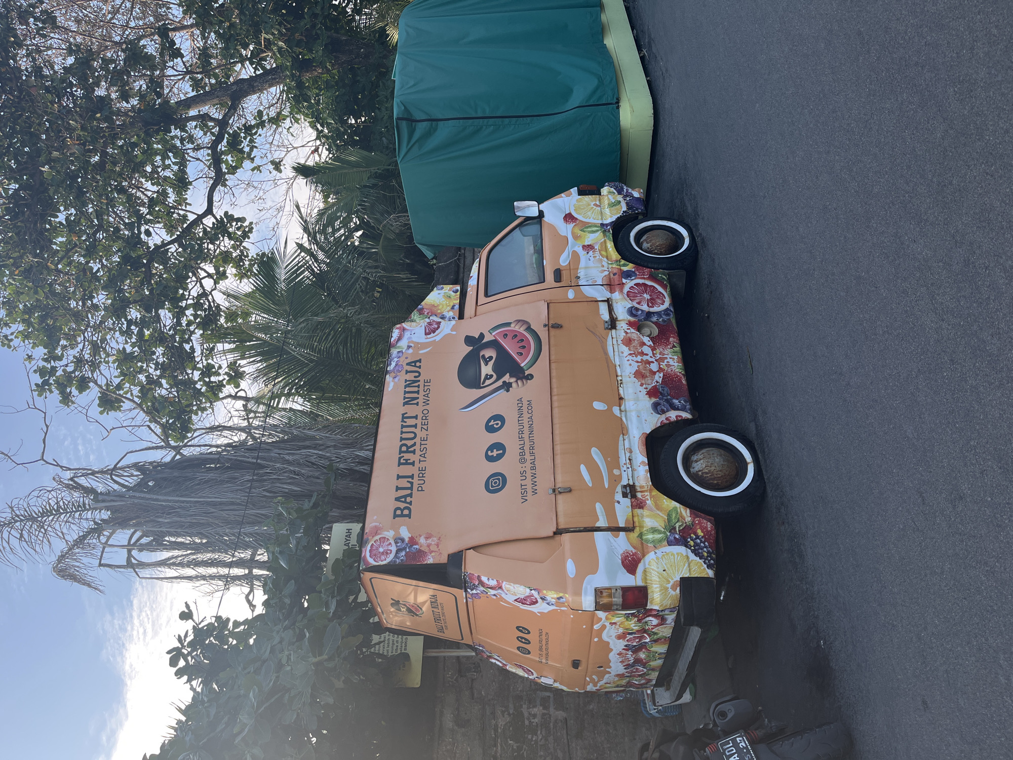 An orange food truck named "BALI FRUIT NINJA" is parked on the side of a street in North Kuta, Indonesia. The truck is adorned with vibrant fruit graphics and features a cartoon ninja character holding a slice of watermelon. Social media handles and a website address are visible on the truck. To the right of the food truck is a large, green-covered structure, possibly a tent or shelter, resting on a yellow base. A motorcycle is partially visible on the left side of the image. The background consists of trees, including a tall, dry palm tree, and a clear blue sky with scattered clouds. The lighting suggests it is daytime.