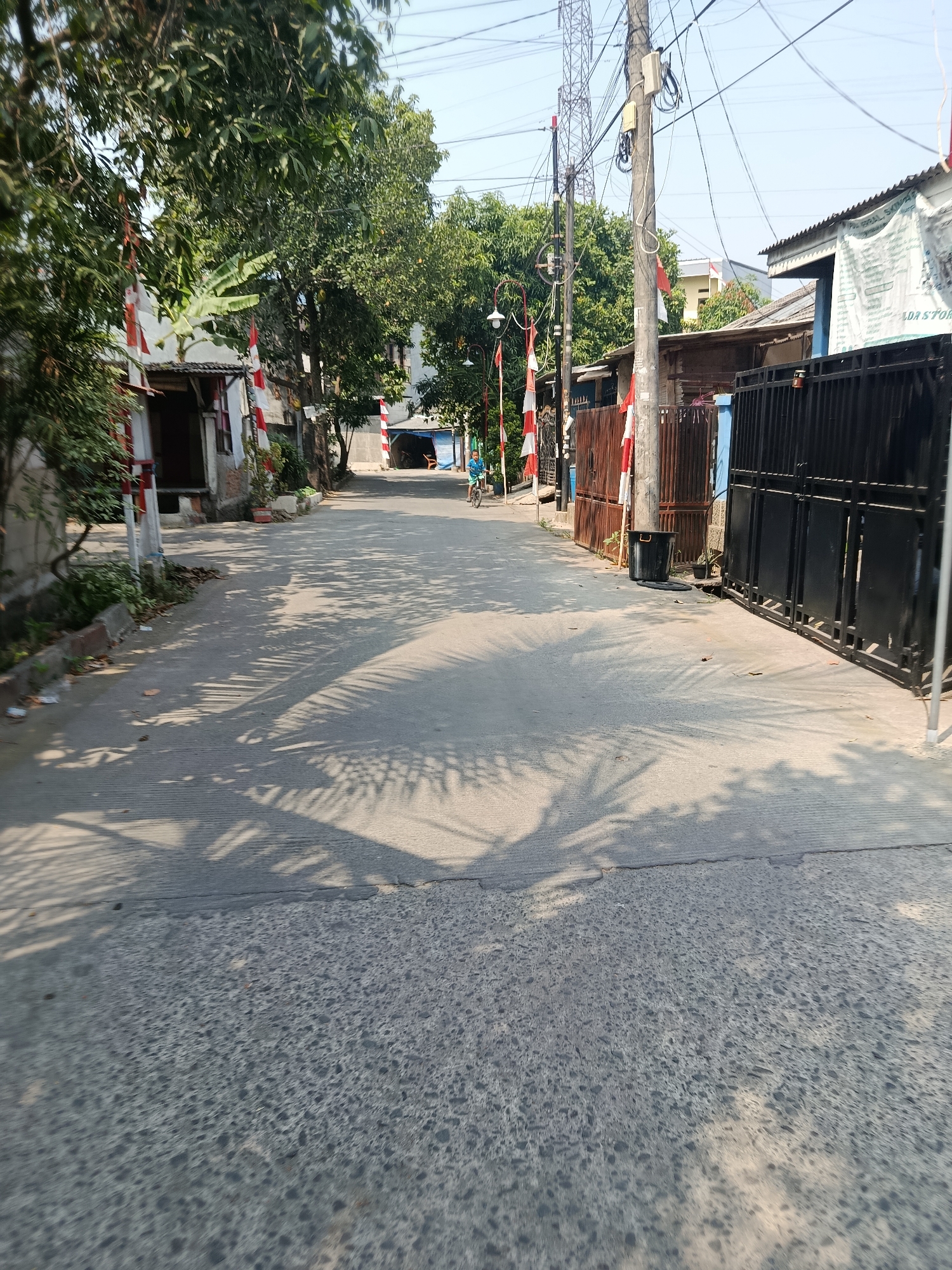 The image shows a residential street with buildings on either side. The street is paved and has palm tree shadows cast across it from the right. There are several utility poles with wires strung between them. Red and white flags are visible, tied to poles along the side of the road, suggesting a festive or commemorative occasion. On the left side of the street, there are trees and a building with a white and red facade. On the right side, there are fences and more buildings. In the distance, a person is riding a bicycle. The sky is clear and blue, indicating a sunny day. The overall impression is that of a moderately populated urban or suburban area, possibly in Southeast Asia, given the presence of palm trees and the style of the buildings.