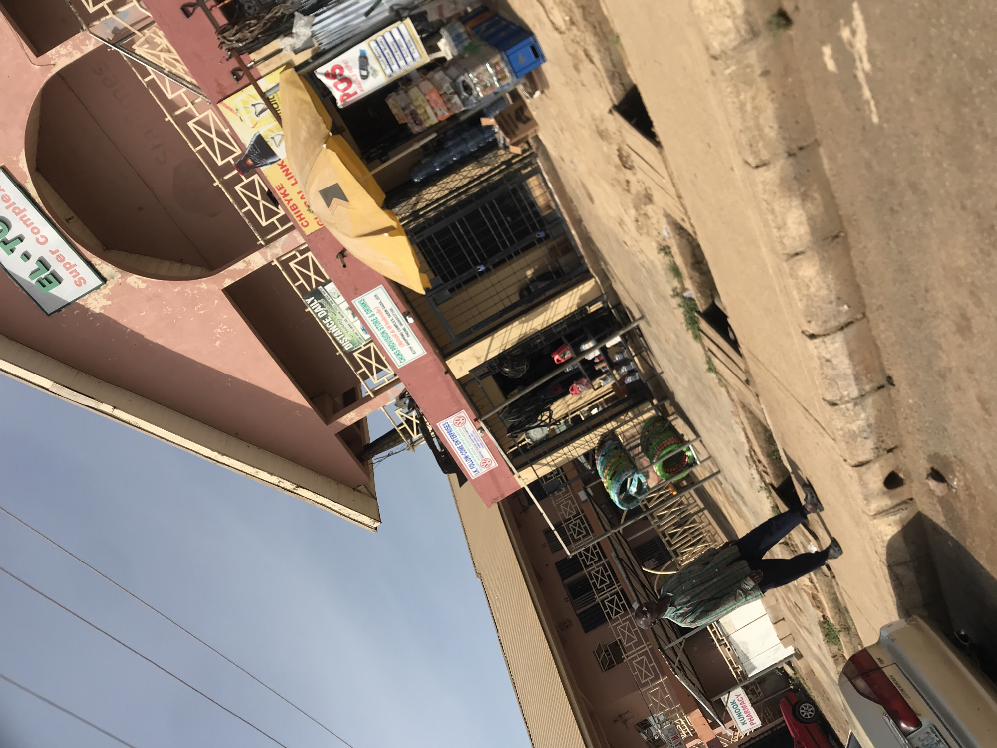 This image depicts a bustling street scene in a commercial district of Jos, Nigeria. The setting features multiple multi-story buildings with light brown or pinkish facades, some including balconies with decorative metal railings. A dirt or unpaved road/sidewalk runs in front of the buildings, with a visible gutter or drainage channel alongside. The sky is clear and light blue, indicating a sunny daytime. Power lines are visible stretching across the upper part of the frame.
One person, wearing a striped long-sleeved shirt and dark trousers, is walking on the sidewalk towards the right side of the image, their back mostly to the viewer. Shops line the street, displaying various merchandise such as bottled drinks, general provisions, and colorful, possibly woven, items on a stand. Parts of parked cars, including a red and a white/beige vehicle, are visible on the right edge of the frame.
The scene is rich with visible text from numerous business signs, offering location cues and details about the establishments. These include "EL-T Super Complex" and "EL-T OF SHOPPING COMPLEX, FARIN GADA, JOS. PHONE 0806953739". Other prominent signs advertise "KUNOOK PHARMACY", "L.K. FOLLOW-COME ENTERPRISES", "DISTANCE DAILY", and "CHUKS PROVISION STORE & DRINKS (Retail & Wholesale)". Further signs read "CHIBYKE GLOBAL LINK", "POS", and "Startimes". The image captures the everyday activity of a vibrant local business area under clear, bright weather conditions.