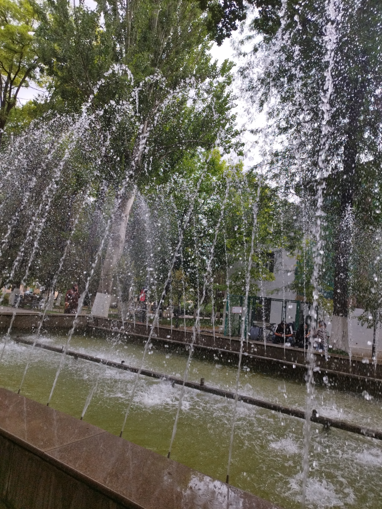 The image depicts a water fountain in a park-like setting, with lush green trees forming the background. Water sprays in vertical streams, creating a curtain of droplets that partially obscures the view of the park. In the mid-ground, several people are visible sitting on benches or walking, suggesting a public recreational area. The water in the fountain basin is a murky green color. The overall scene suggests an outdoor public space, possibly a park or plaza. The location is identified as being in the city of Shymkent, Kazakhstan.