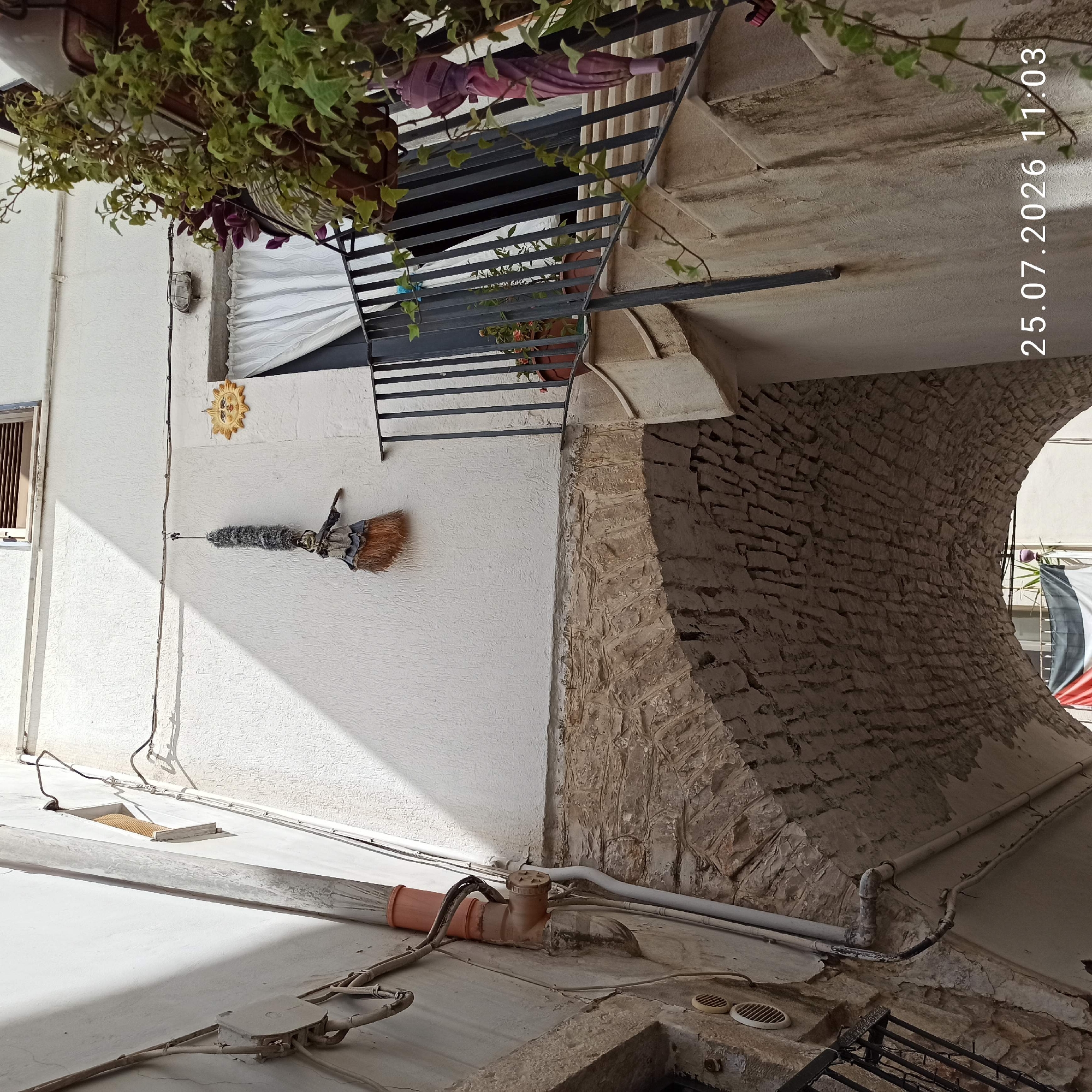 A low-angle, outdoor, medium shot captures a narrow street in Ruvo di Puglia, Italy, on a sunny day. The scene is dominated by the rough-textured white stucco walls of buildings flanking the street. An arched stone passage, likely part of an old building, spans the width of the lower portion of the frame. Above the arch, a rustic broom with a wooden handle and a grey, brush-like head is suspended from a wire across the wall. To the right of the broom, a decorative yellow ceramic sun with a human face is affixed to the wall. Further to the right, a dark metal railing and steps lead up to a balcony where potted plants, including an ivy, are visible, along with a closed pink umbrella hanging from the railing. Electrical conduits and pipes are visible along the building walls. A date stamp "25.07.2026 11:03" appears in the lower right corner.