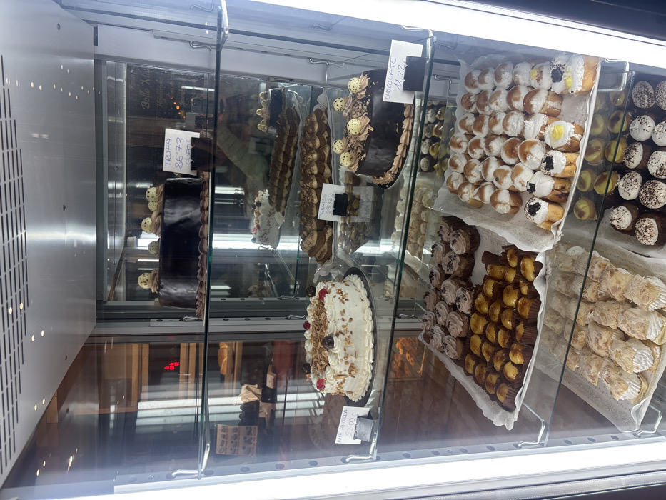 This image depicts a brightly illuminated, multi-tiered glass display refrigerator filled with various baked goods and confections inside a pastry shop or bakery in Salamanca, Spain.
The top shelf features a large, dark chocolate cake adorned with several light-colored spheres and chocolate shavings, priced at "TRUFA 26'73 €/Kg". Reflections on the glass hint at a shop name, possibly "Bello Monte."
The second shelf (from top) holds three distinct offerings:
* On the left, a round white cake topped with chopped nuts and a few red berries, labeled "Tarta Americana 22,00 €".
* In the center, a tray of numerous individually portioned items resembling tiramisu, priced at "TIRAMISÚ 23,32 €".
* On the right, a smaller, dark chocolate cake, also decorated with light spheres and chocolate shavings, labeled "CHOCOLATE 11,22 €".
The third shelf displays multiple trays of smaller, individual pastries:
* On the left, a tray of uniformly sized, square-shaped mini pastries in dark paper cups, appearing to be mini cupcakes or similar.
* In the center, a large tray contains a variety of small, round, and elongated individual pastries, some topped with cream, fruit, or chocolate.
* On the right, another tray holds numerous mini cupcakes with light frosting and chocolate chip toppings.
The bottom shelf contains more trays of pastries:
* On the left, elongated pastries with a flaky, almond-dusted top.
* On the right, dark-colored mini cupcakes topped with white frosting and dark sprinkles.
No people or direct interactions are visible. The environment is the interior of the refrigerated display case itself.