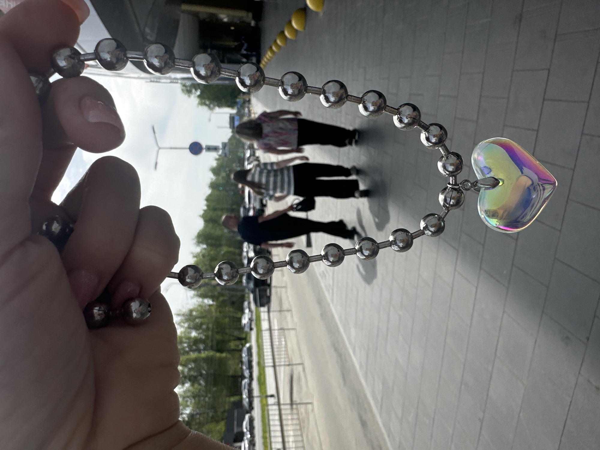 The photograph is taken in Nizhny Novgorod, Russia. A hand holds a silver ball chain with an iridescent heart pendant, partially obscuring the background. Three individuals walk in the background: the first wears a light pink floral top, the second a striped shirt, and the third a navy shirt. Cars are parked on the left behind a low fence, and the sidewalk is to the right. Overhead, yellow barrier bollards border a building.