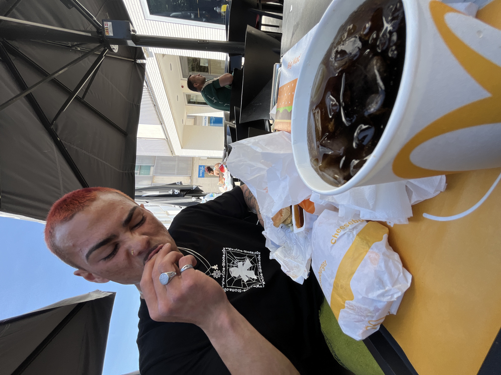 Here's a description of the image:
The photograph shows a young man with a reddish-orange buzzcut, eating outside at a table in what appears to be a McDonald's in Barcelona, Spain. He is wearing a black t-shirt with a graphic design on the front. He has a silver ring on each hand and a nose ring. He is in the act of eating, with his eyes closed slightly.
On the table, there is a large cup of cola with ice, a cheeseburger wrapped in paper, and other food containers with napkins. The table has a yellow and black design on it.
The location is an outdoor patio, shaded by a black umbrella. In the background, buildings typical of Barcelona architecture can be seen, and another man is seated at a table.
