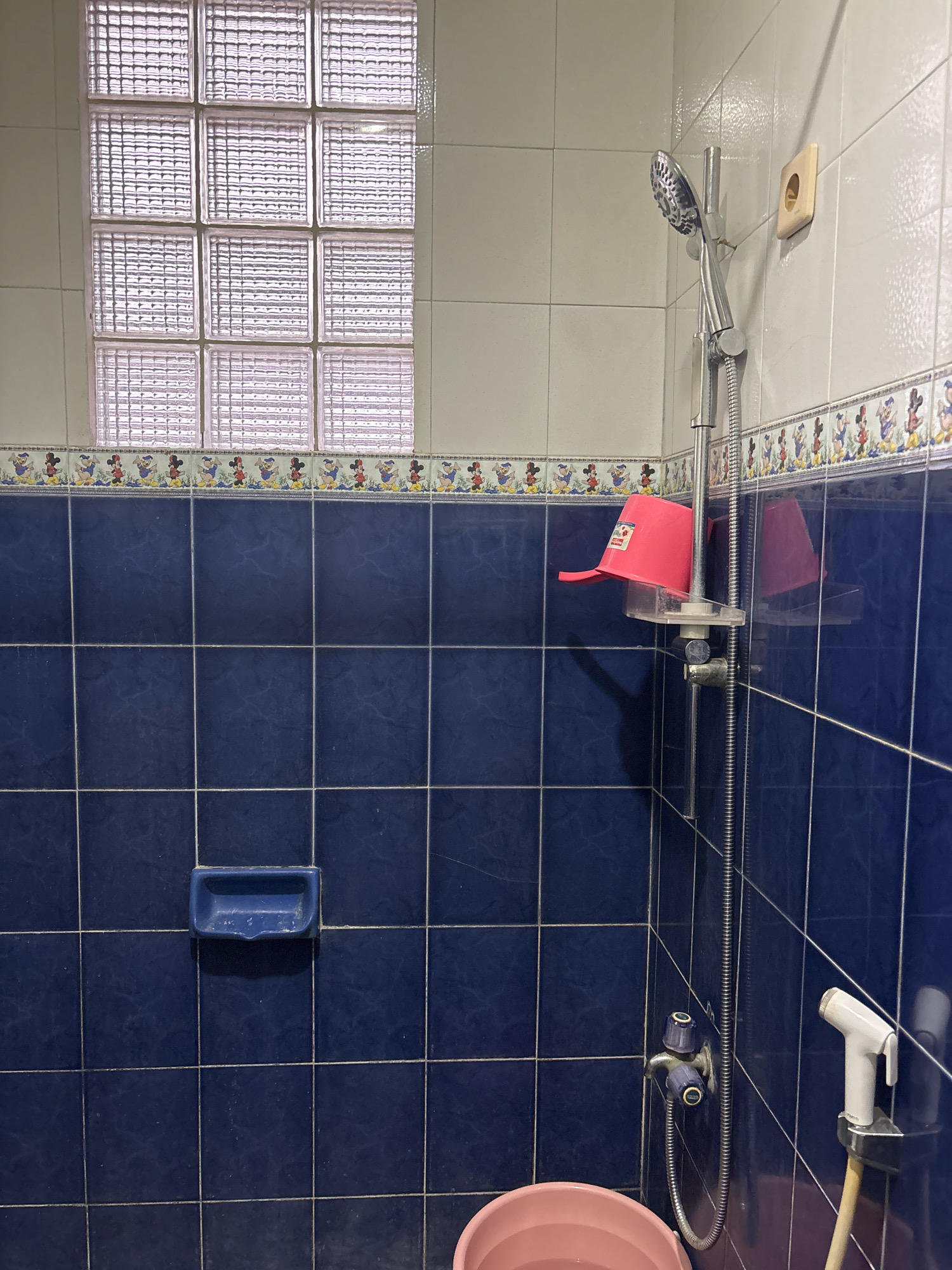 The image depicts a small, tiled bathroom in a residential setting, likely in Bekasi, Indonesia. The walls are covered in dark blue square tiles, with a decorative border featuring cartoon characters above the blue tiles. At the top of the wall is a window made of frosted glass blocks.
A shower head is mounted on the wall, attached to a flexible hose. Below the shower head, a pink plastic bucket is hanging from a clear plastic holder. To the left of the shower, a blue plastic soap dish is affixed to the blue tiles.
Further down the wall, a faucet with two blue knobs controls the water flow for the shower. A white bidet sprayer is also visible on the right side of the shower area. In the foreground, the rim of a pink plastic basin or bucket is visible. The overall impression is of a functional, albeit somewhat dated, bathroom.