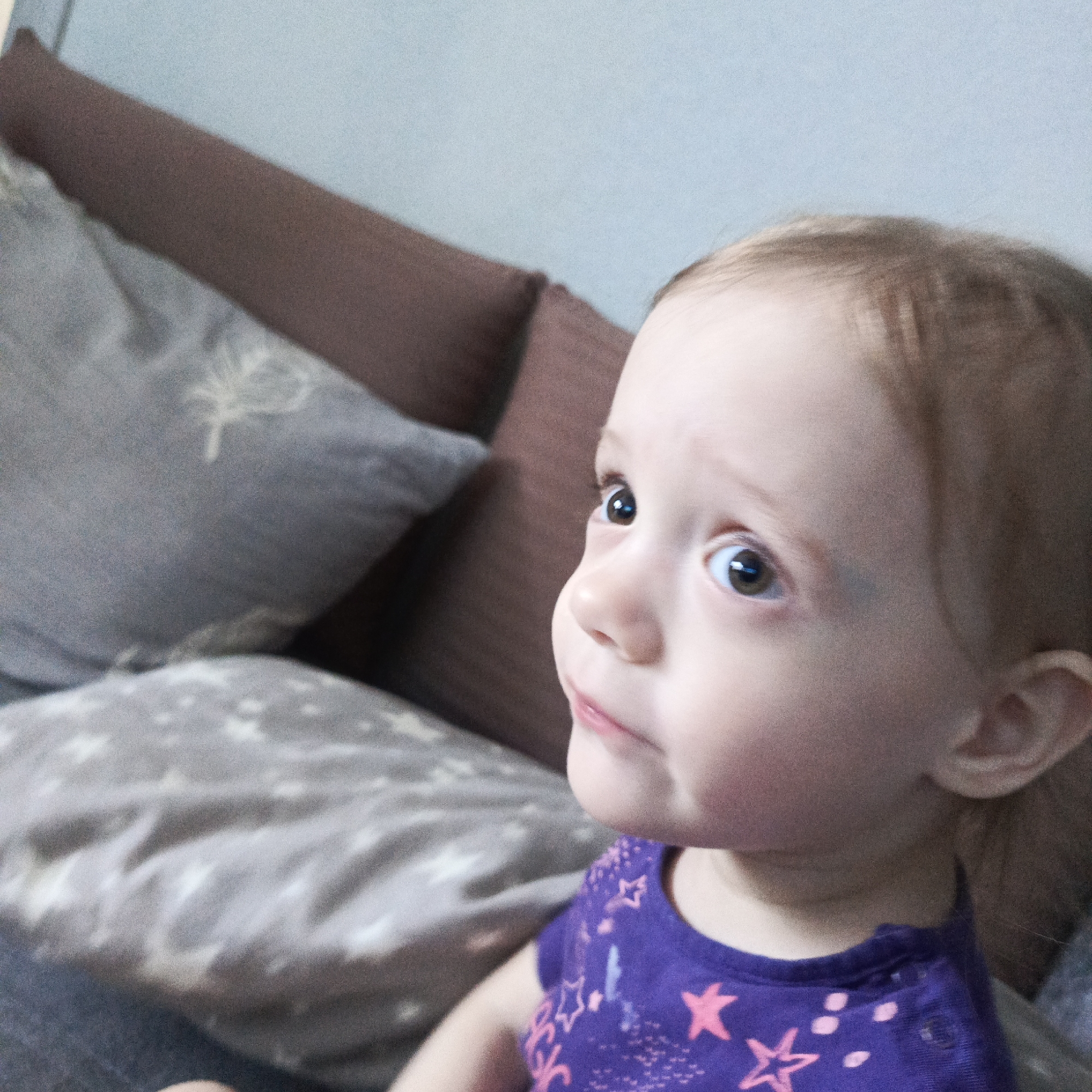 The image shows a young child with light skin, brown hair, and brown eyes looking up and to the side. She is wearing a purple shirt with pink star patterns. She appears to be sitting on a sofa or bed with several pillows behind her. The pillows are gray with patterns and a solid brown. The location is unknown and there is no evidence to support the location being in Марковский, Russia.