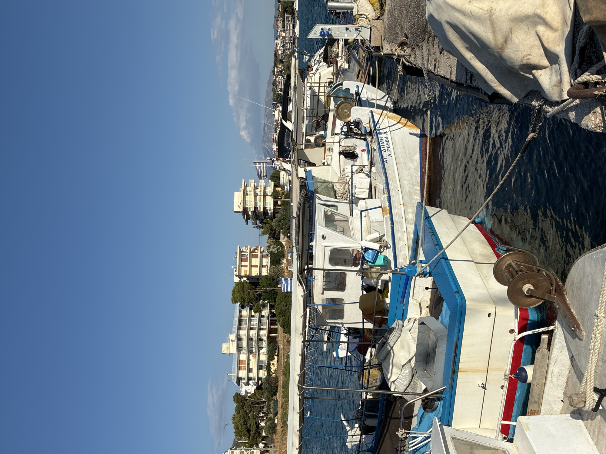 In Limenas Markopoulou, Greece, several fishing boats are docked in a harbor under a clear blue sky. The boats are white with blue trim and some have blue canopies. One boat in the foreground has the Greek lettering "ΑΓ. ΔΗΜΗΤΡΙΟΣ Λ.Ρ.6584 A" visible on its side. In the background, there are multi-story buildings with balconies and trees. The water in the harbor is calm and dark blue. The scene appears to be a typical day in a Greek fishing village.