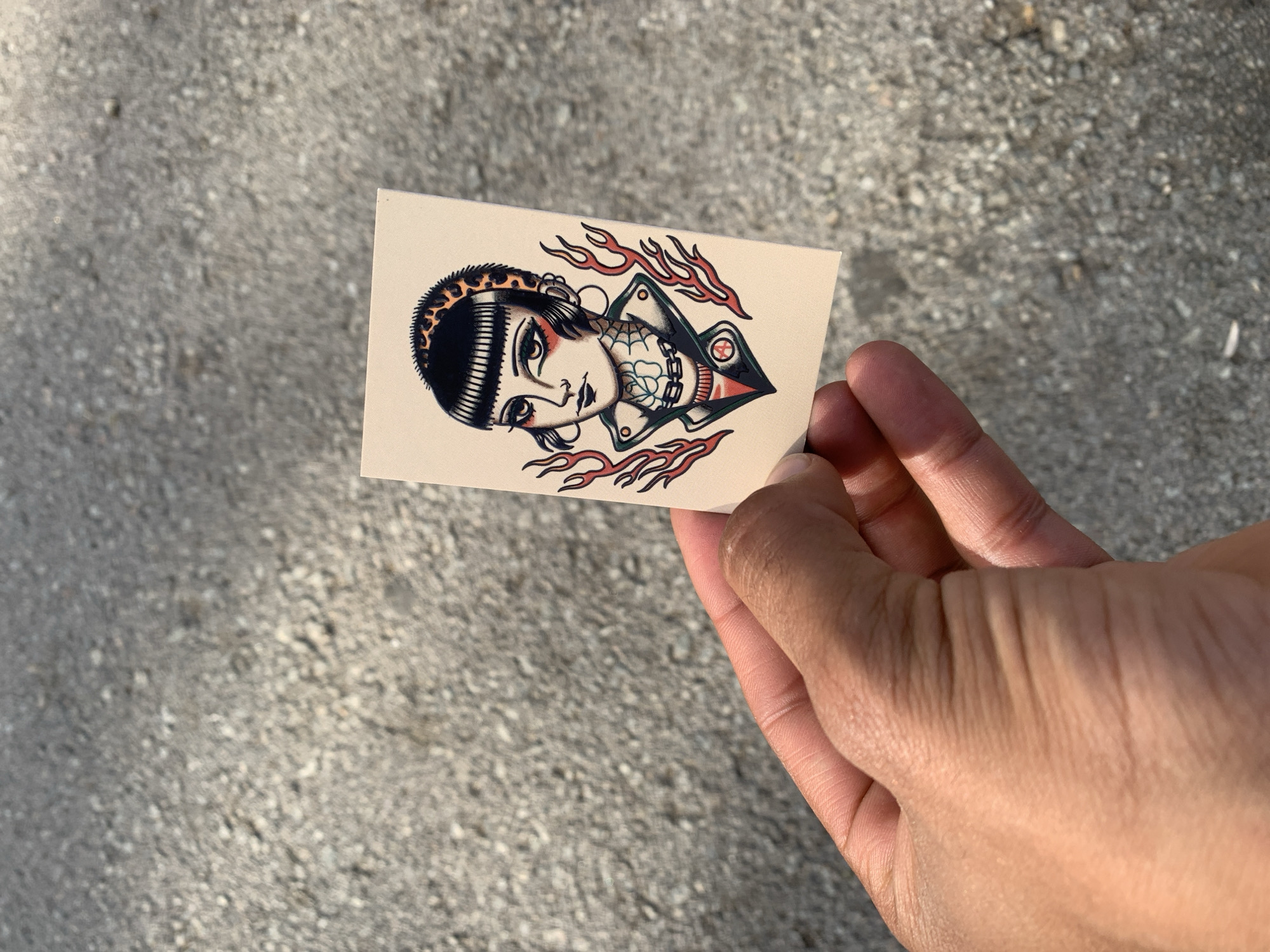A hand holds a rectangular card with a tattoo-style illustration of a woman's face and shoulders. The woman has short black hair with a leopard print headband, heavy eyeliner, and hoop earrings. She is wearing a leather jacket over a red shirt and has a chain necklace. Flames are depicted on either side of her. The card is placed against a blurred background of grey gravel. There is no indication that this image was taken in Barcelona, Spain.