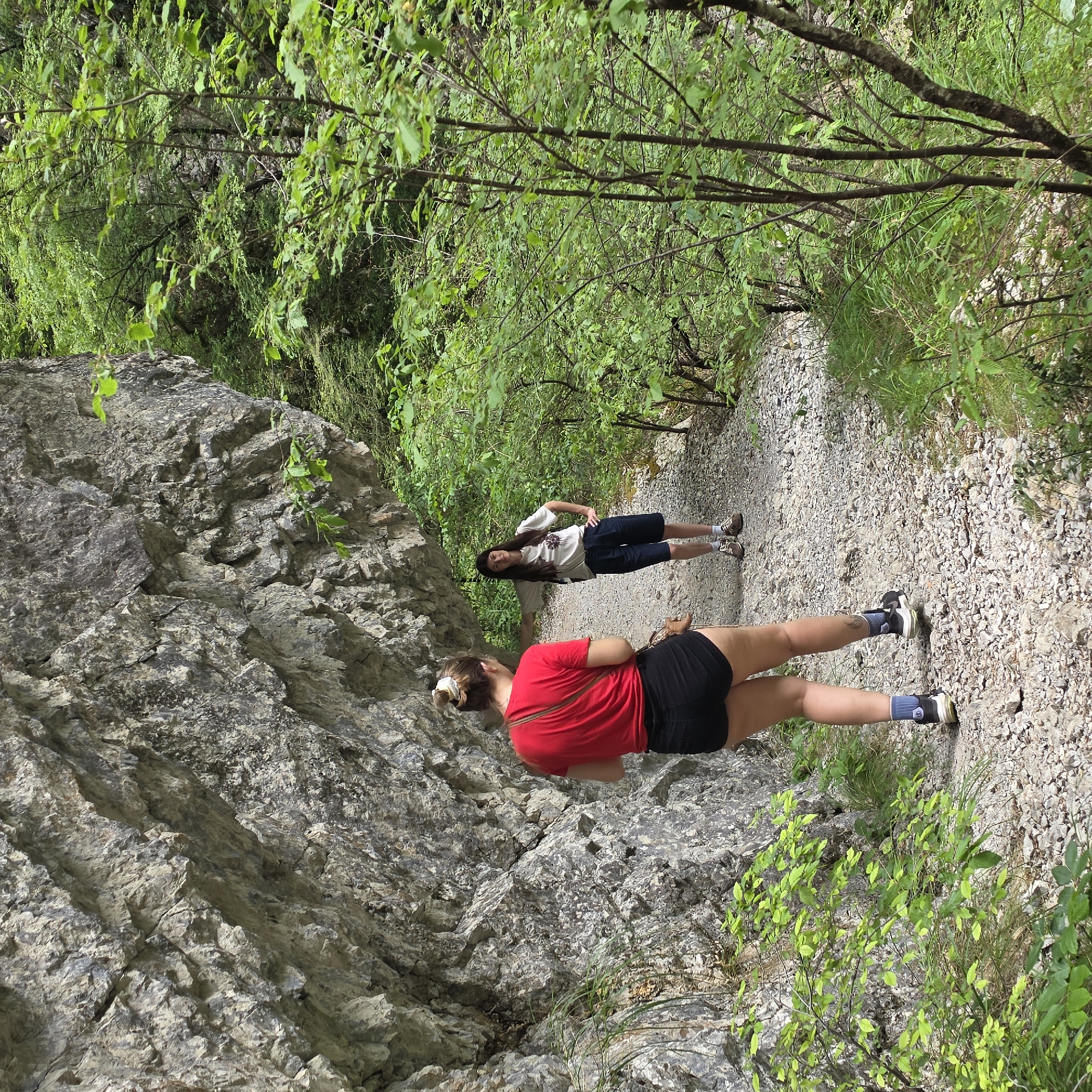The image shows two women standing on a gravel path in a wooded area in Lazba, Montenegro. The path is surrounded by green trees and foliage. To the left of the path is a large rock formation. The woman in the foreground is wearing a red shirt and black shorts. The woman in the background is wearing a white shirt and dark shorts.
