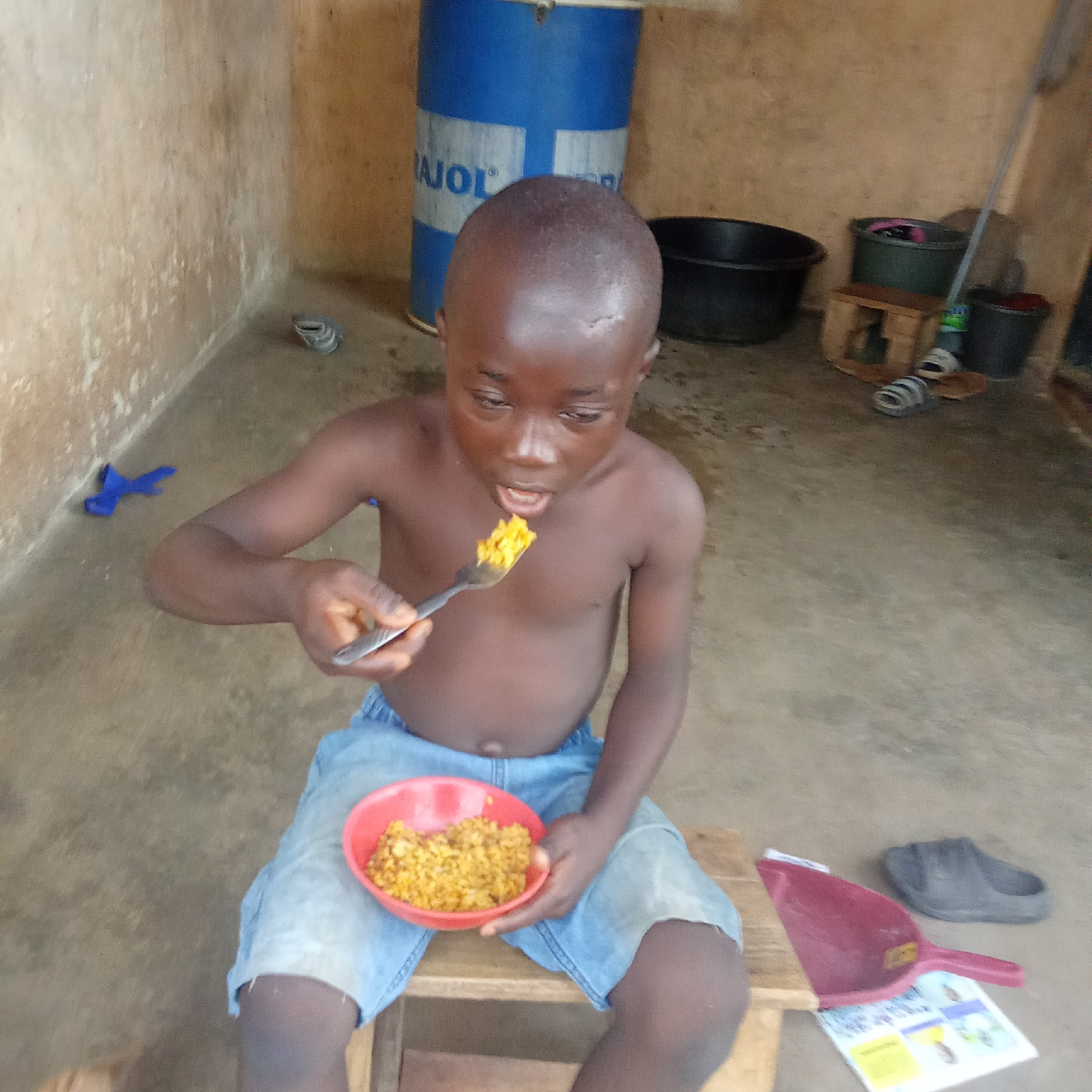 A young Black boy, appearing to be in early childhood, is depicted in a full-shot, seated on a small wooden stool and eating a meal. He is shirtless and wears light blue denim shorts. He holds a metal spoon with a portion of yellowish-orange rice or a grain-based meal towards his mouth, while a red plastic bowl filled with the same food rests on his lap. His expression is focused and neutral, concentrated on eating.
The setting is a simple, rustic indoor or sheltered outdoor area, likely within a home or utility space in Akwanga, Nigeria. The walls are light brown, possibly concrete or mud, showing some wear and patches of white. The floor is a light grey concrete or packed earth.
In the background, a large blue metal barrel with partially visible white text, reading "AJOL", stands upright. Various household items are scattered around the space: a black plastic basin, a dark green bucket, another dark bucket, and a mop leaning against the wall. Several pairs of sandals or slippers, including a grey rolled-up pair and a black pair, are on the floor. A red plastic dustpan and a white booklet or piece of paper with illustrations are also visible on the ground. The lighting suggests it is daytime.