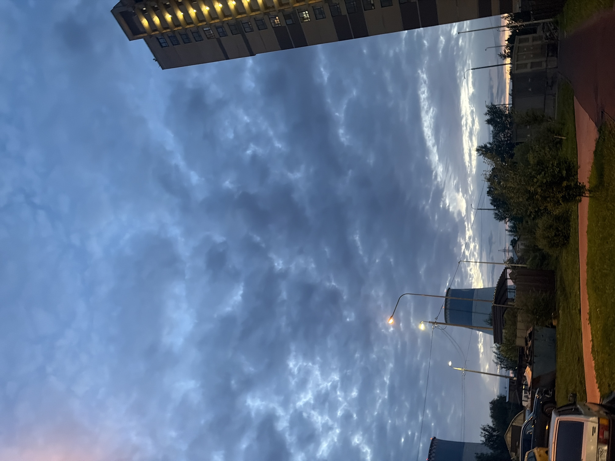 The image shows an outdoor scene at dusk or dawn, likely in an urban or industrial area of Saint Petersburg, Russia. The sky is filled with dark, cloudy formations, with patches of lighter sky peeking through, suggesting the presence of twilight. In the foreground and mid-ground, there are several objects. To the right, a tall, modern apartment building is visible, with illuminated windows that create a pattern of yellow lights on its facade. In the center and left of the image, there are large, cylindrical structures that resemble cooling towers, common in industrial facilities. There are also streetlights, some illuminated. Several parked cars and what appear to be discarded materials or storage containers are scattered around the ground. Vegetation, including trees and grass, is also present, indicating some green space amidst the industrial and residential elements. The overall atmosphere is somber and somewhat industrial, characteristic of many areas within large cities.