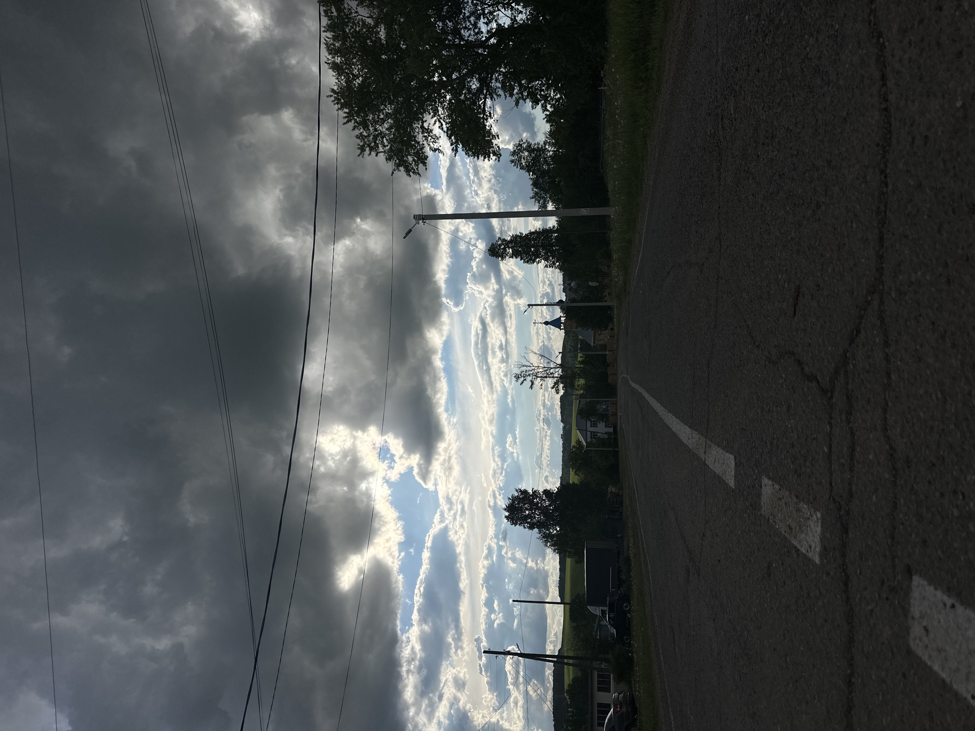 The image shows a view along a paved road with a broken white line marking the lane. The road appears to lead towards the horizon, where there is a visible structure with a blue dome, possibly a church or religious building. Power lines run parallel to the road on the left side of the frame. The sky is divided between dark, stormy clouds on the left and patches of bright, sunlit clouds on the right. Trees and greenery are visible alongside the road, suggesting a rural or suburban setting. Based on the location information, the scene is likely in or near Альняш, Russia. The image contains no visible people or actions, focusing solely on the landscape and infrastructure.