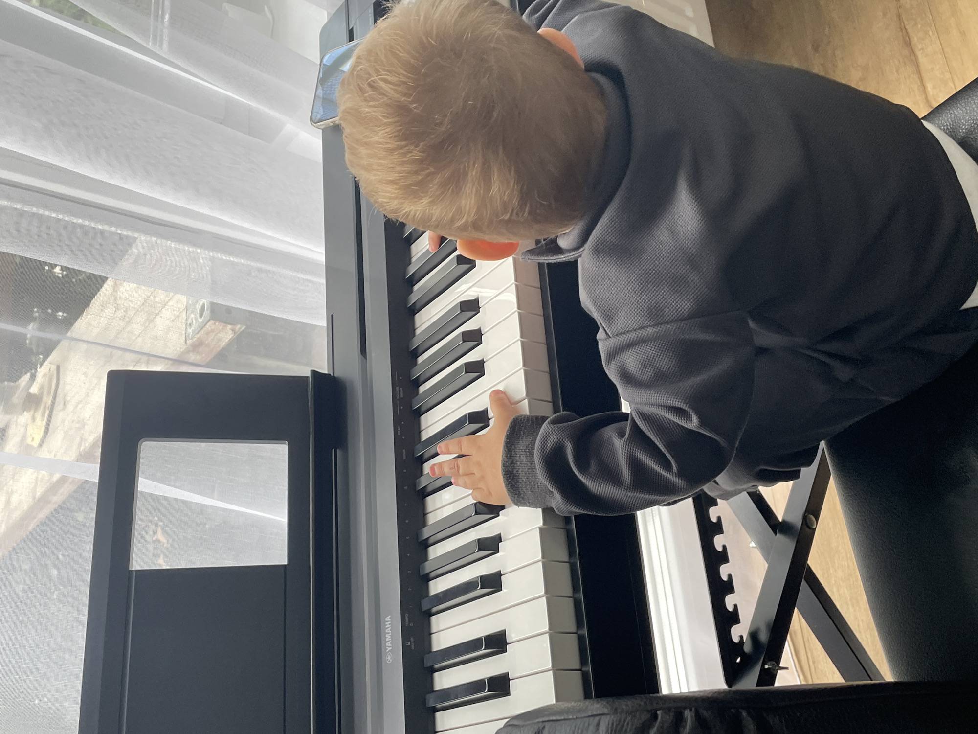 A young child, viewed from behind, is seated at a Yamaha digital piano, pressing keys with their right hand. The child is wearing a dark grey long-sleeved shirt and is positioned in front of a window with sheer white curtains. A black music stand is attached to the top of the piano, with a small rectangular opening revealing the view outside. The piano is placed on a black, X-shaped stand, and the child is sitting on what appears to be a black leather chair. A smartphone is placed on the piano to the right of the child's head. The background suggests an indoor domestic setting with natural light filtering through the window. The image does not contain information to determine its location in the city of Ulmen, Germany.