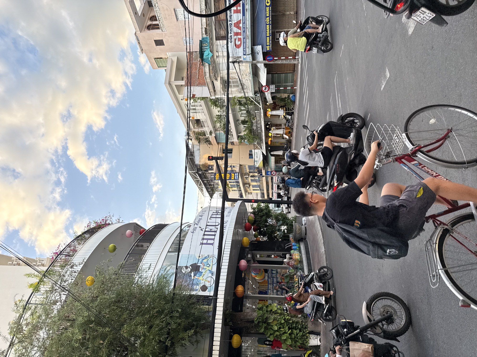 The image depicts a busy street intersection in Nha Trang, Vietnam, during daylight. Numerous motorbikes navigate the roadway, with a cyclist in the foreground. Buildings line the street, displaying various signs in Vietnamese, including "CUA HANG PHU K." and "BON MUA COFFEE." Some buildings feature balconies adorned with colorful lanterns. The sky is partly cloudy, with a mix of blue sky and scattered white and yellow clouds. People on motorbikes are dressed casually, some wearing helmets. The overall scene captures the vibrant atmosphere of a city street in Southeast Asia.