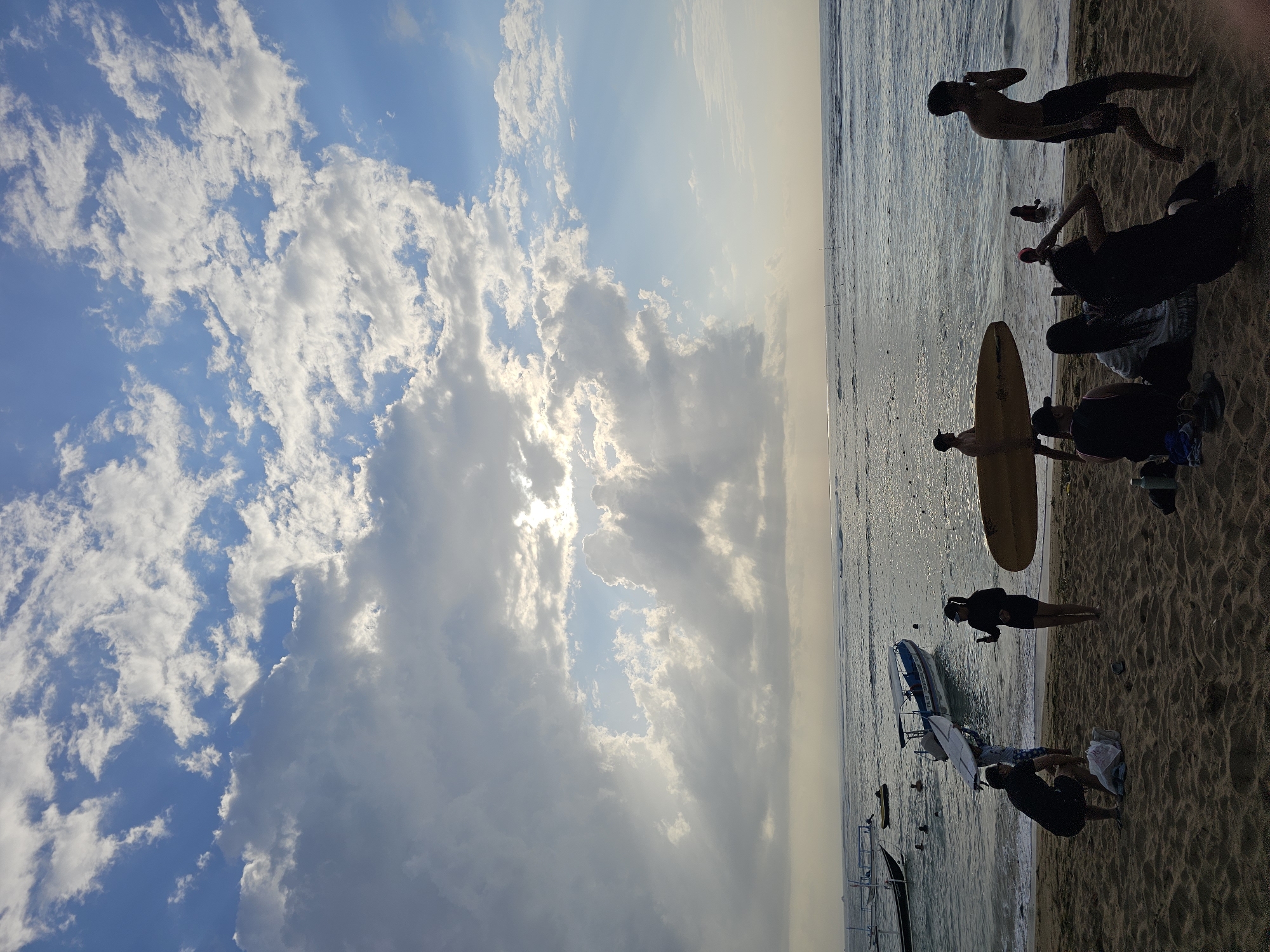 The image depicts a beach scene at what appears to be Sanur Beach in Denpasar, Indonesia, during the late afternoon or early evening. The sky is filled with dramatic clouds, with the sun partially obscured, creating visible sunbeams filtering through. The ocean is calm, with gentle waves lapping the shore. Several people are present on the beach and in the water.
In the foreground, on the sandy beach, a group of people are gathered. One person is kneeling, possibly unpacking items from a bag. Another person is standing, holding a phone, as if taking a picture. Further to the right, a person is walking along the beach, shirtless and wearing shorts, with their hand near their face. There are also several individuals sitting on the sand, some with their backs to the camera.
In the water, several people are swimming or wading. A yellow surfboard is propped up near the shore, and another person is carrying a surfboard. Several boats are anchored or moored in the shallow water, including a larger boat with a canopy and smaller, traditional fishing boats.
The overall atmosphere suggests a relaxed, recreational setting, likely a popular spot for both locals and tourists to enjoy the beach and water activities. The lighting indicates either sunrise or sunset, contributing to the picturesque quality of the scene.