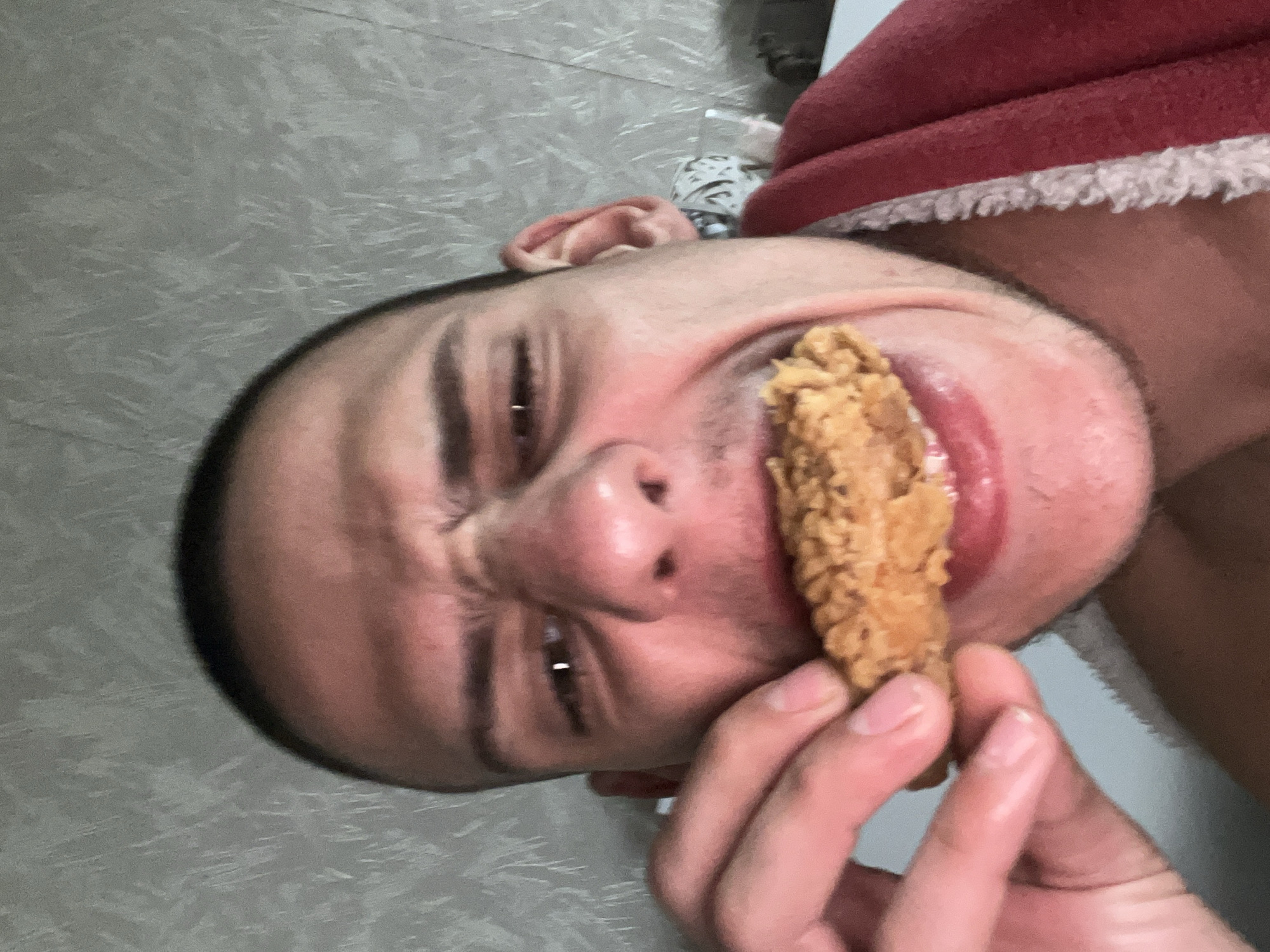 The image, captured in Sofia, Bulgaria, presents a close-up, tilted portrait of a person, seemingly male, against a grey, textured background. The subject has short dark hair and a light skin tone. He is engaged in the act of eating, holding a golden-brown piece of fried chicken (appearing to be a drumstick or wing) up to his open mouth with his left hand.
His facial expression conveys a mix of intense concentration and anticipation, with his eyes looking slightly upwards and to the side, and distinct wrinkles visible around his eyes and nose. On his upper right side, a red garment with white, fuzzy trim is visible, suggesting a festive item like a Santa hat or robe. The background features a consistent grey tone with a subtle, abstract pattern, possibly an indoor floor or wall. The lighting is uniform, indicating an indoor setting. No text is visible in the image.