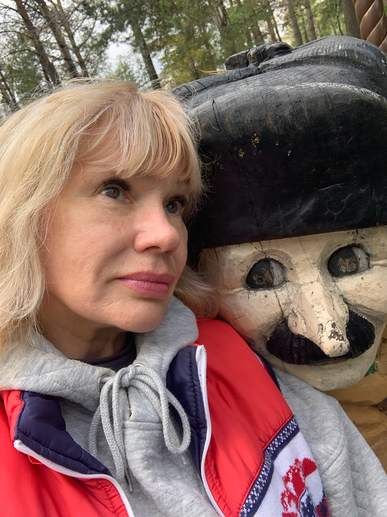 A woman with blonde hair and a red, white, and blue jacket stands next to a weathered wooden figure of a man with a black hat and a mustache. The background shows trees and a cloudy sky. The image appears to be a selfie taken outdoors, possibly in a park or wooded area. The wooden figure's face is cracked and worn, suggesting it is an older decoration or statue. The woman is looking off to the side, and the wooden figure is positioned behind her, its head tilted slightly.