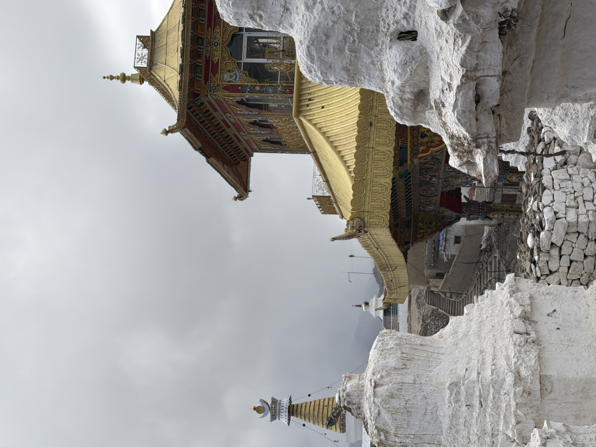 In the city of Leh, India, a Buddhist monastery complex sits under a cloudy sky. The main subject is a multi-tiered golden roofed building with ornate red and gold trim. To the left, a tall stupa with a golden spire and a crescent moon finial stands, also yellow and white. In the foreground, two weathered white structures, resembling broken stupas, are visible. A stone pathway with steps leads up towards the monastery in the background, with other smaller white stupas and buildings scattered throughout the complex. The ground is covered with rubble and stones.