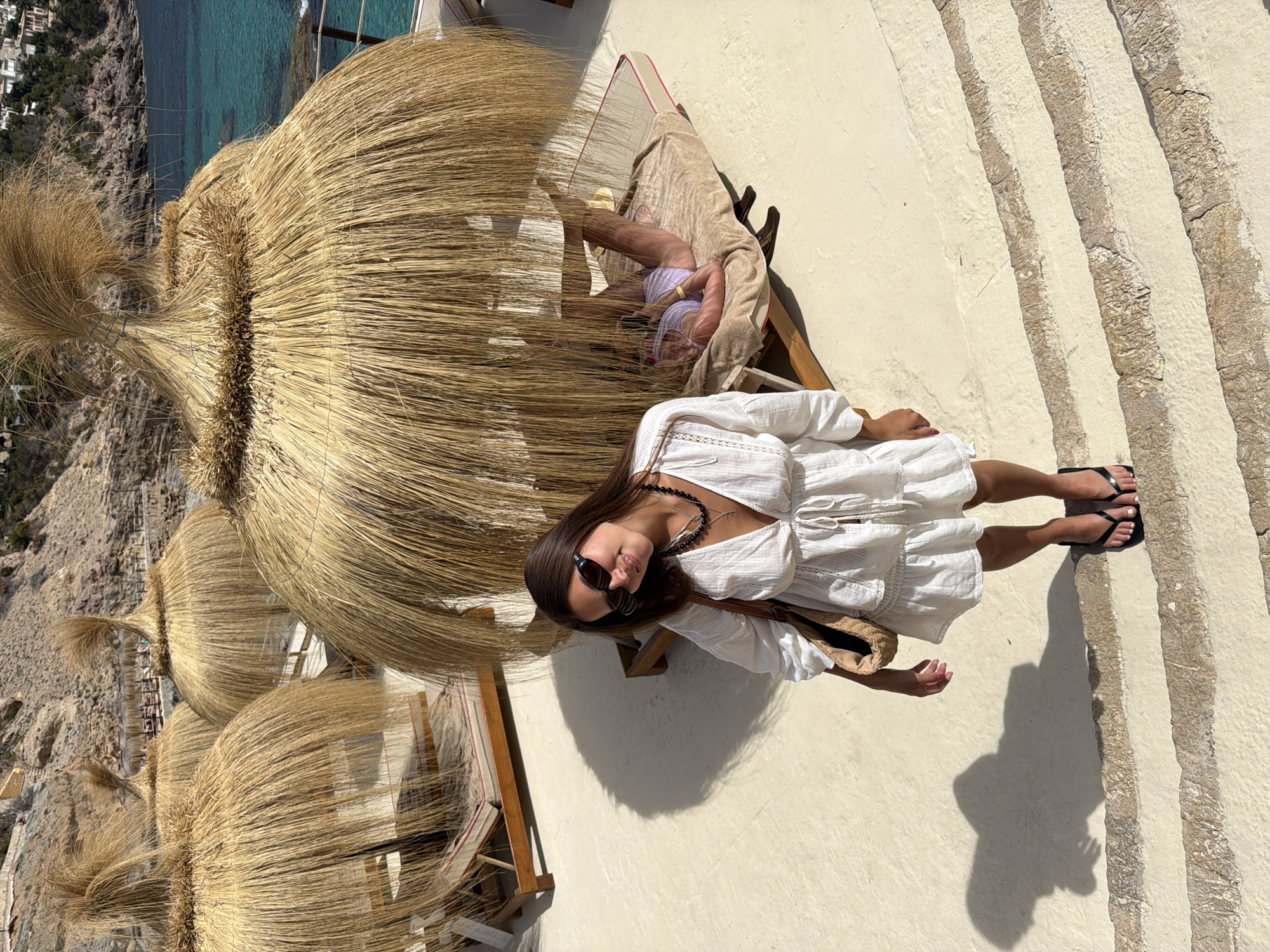 The image depicts a woman standing on a stone terrace in Port D'andratx, Spain. She is wearing a white dress, black sunglasses, and black flip-flops. She is also carrying a straw bag. In the background, there are several thatched umbrellas, lounge chairs, and the Mediterranean Sea. The woman is smiling and looking at the camera. Another person is visible lying on a lounge chair under one of the umbrellas. The scene appears to be a resort or beach club.
