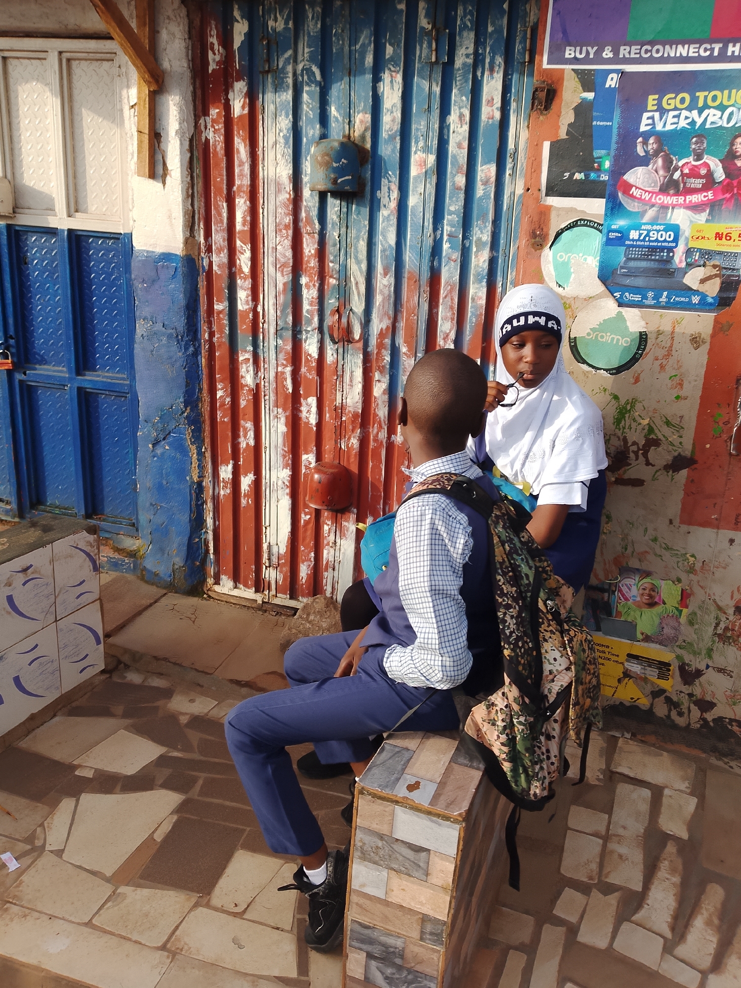 Two children, a boy and a girl, are sitting on a stone bench in front of a corrugated metal building with red and blue paint in Gura Top, Nigeria. The boy, wearing a white and blue checkered shirt and blue pants, is looking at the girl and has a backpack on. The girl, wearing a white hijab and blue uniform, is looking down at her hands and holding a pair of glasses. The background shows a blue metal door and a colorful advertisement poster on the wall.