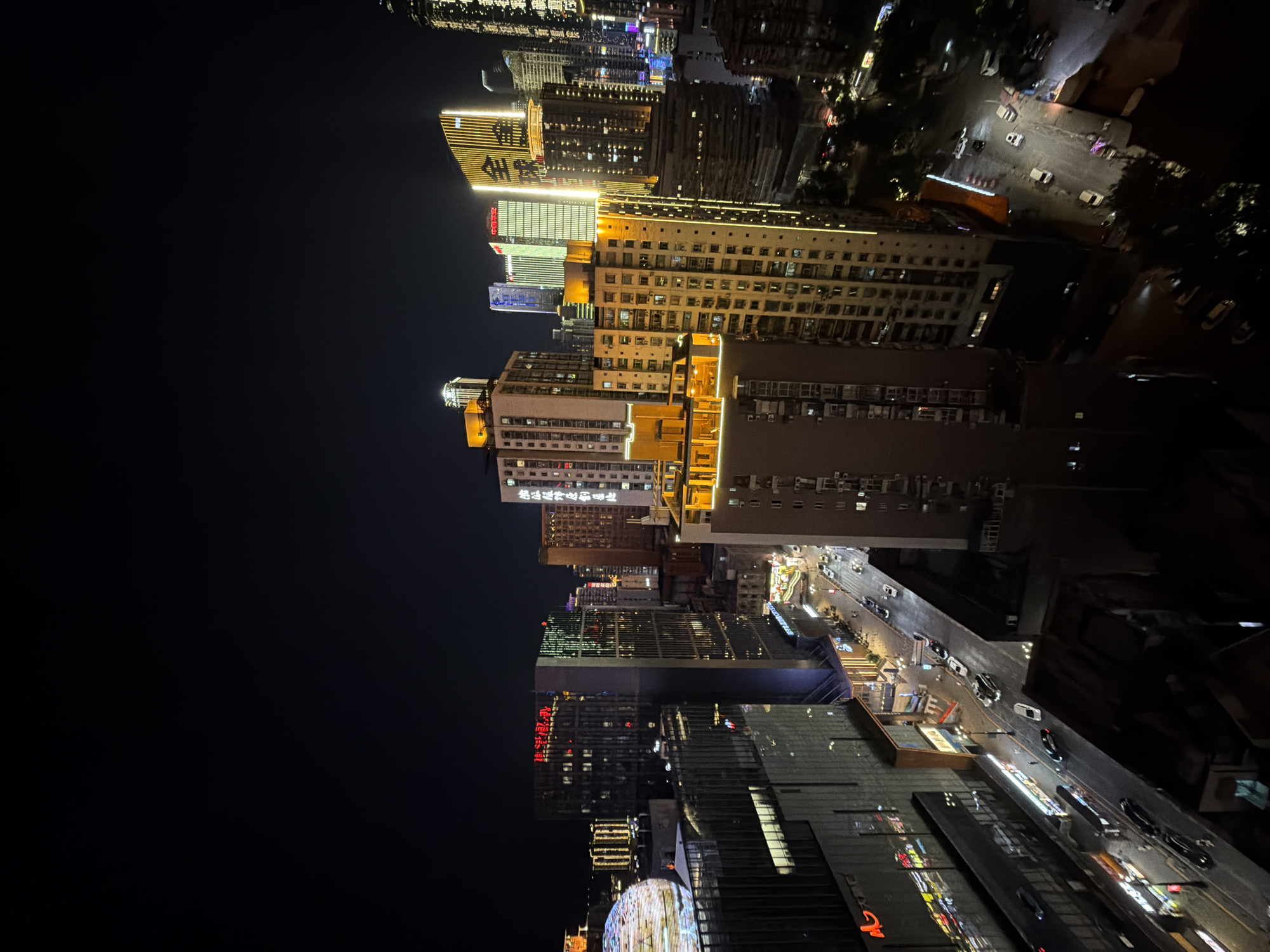 This is a nighttime aerial view of a dense urban cityscape in Yuzhong District, China, characterized by a multitude of illuminated skyscrapers and buildings. The foreground features a dark, shadowed area, likely the roof or edge of a lower building. To the left, a large building with a dark facade and illuminated windows is visible, with a rounded, partially lit structure atop it, resembling a dome or sphere adorned with a pattern of lights.
Dominating the center and right of the image are various tall buildings of different architectural styles, many of which are brightly lit. Some buildings have vertical light strips in yellow and white, outlining their structures. One prominent building in the center, with a light yellow facade, has rows of yellow lights outlining its horizontal sections and vertical edges. To its right, another tall building features a facade covered in vertical yellow lights, creating a striking illuminated pattern. Further right, a more traditional skyscraper with numerous illuminated windows rises into the dark sky, its silhouette defined by its lit panes.
Below the buildings, a street runs through the scene, illuminated by streetlights and the glowing signs of businesses. Several cars are visible on the street, their headlights and taillights creating streaks of light, indicating movement. A few pedestrians can be seen on the sidewalks, though they are small and indistinct due to the distance and lighting conditions. The overall impression is of a vibrant, active city at night, with a strong emphasis on architectural lighting and the energy of urban life.