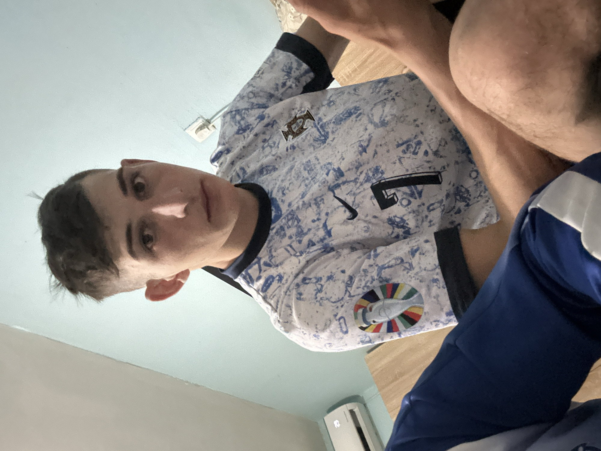 A young man is positioned in the frame, wearing a white and blue patterned soccer jersey with the Portuguese emblem, number "7", and the UEFA Euro 2020 patch. He is seated, with what appears to be his leg or thigh visible on the right side of the frame. The background shows a light blue wall and a portion of what seems to be an air conditioning unit. The image is taken from a low angle. There is no information to confirm this photo was taken in Tsarevo, Bulgaria.