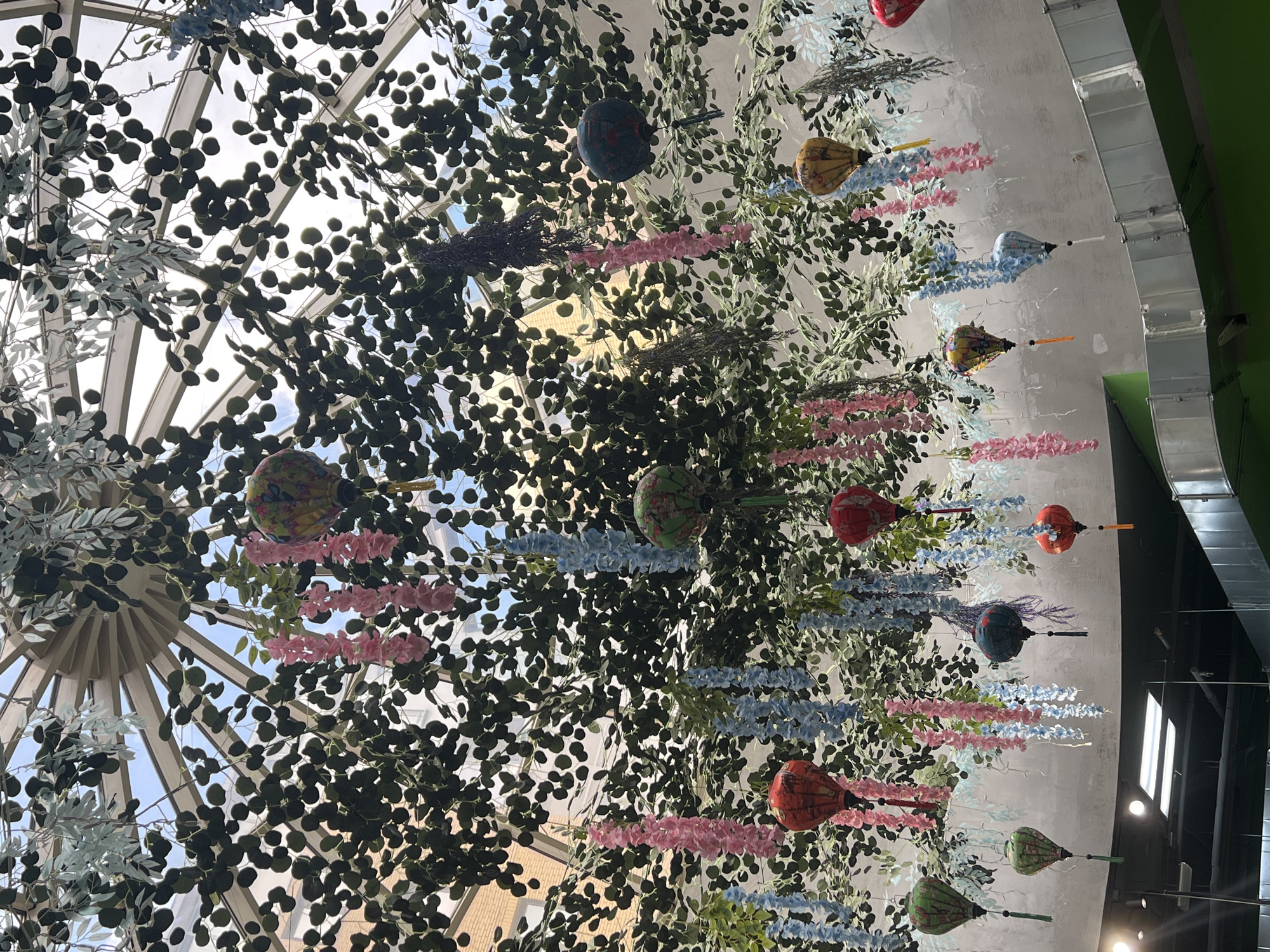 The image depicts an interior view of a structure with a glass dome ceiling, possibly a conservatory or atrium. Through the glass, hints of the sky can be seen. Dense foliage and decorative elements, including lanterns and hanging floral garlands, are suspended from the ceiling. The decorations are predominantly pink and blue. Ductwork runs along the interior architecture. The specific location is Вёшки, Russia.