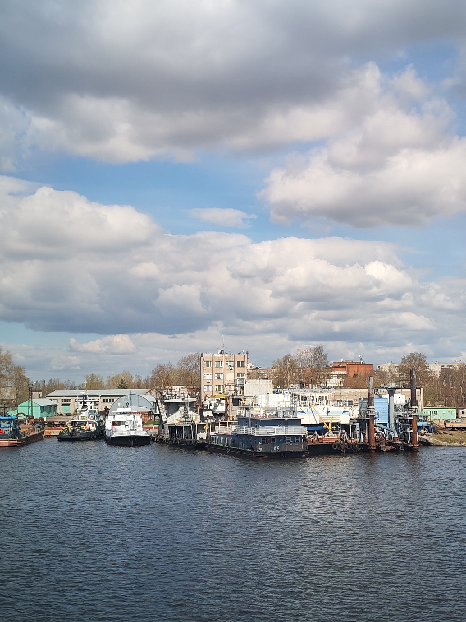 The image shows a waterfront scene in Cherepovets, Russia. Several boats and barges are docked along the shoreline of a dark body of water, likely a river or lake. Buildings of varying sizes and styles are visible on the shore behind the docks, including what appears to be a multi-story residential or office building. The sky is partly cloudy, with patches of blue sky interspersed among large, white and gray clouds. The scene is shot in daylight, with a clear view of the docks and buildings along the water's edge.