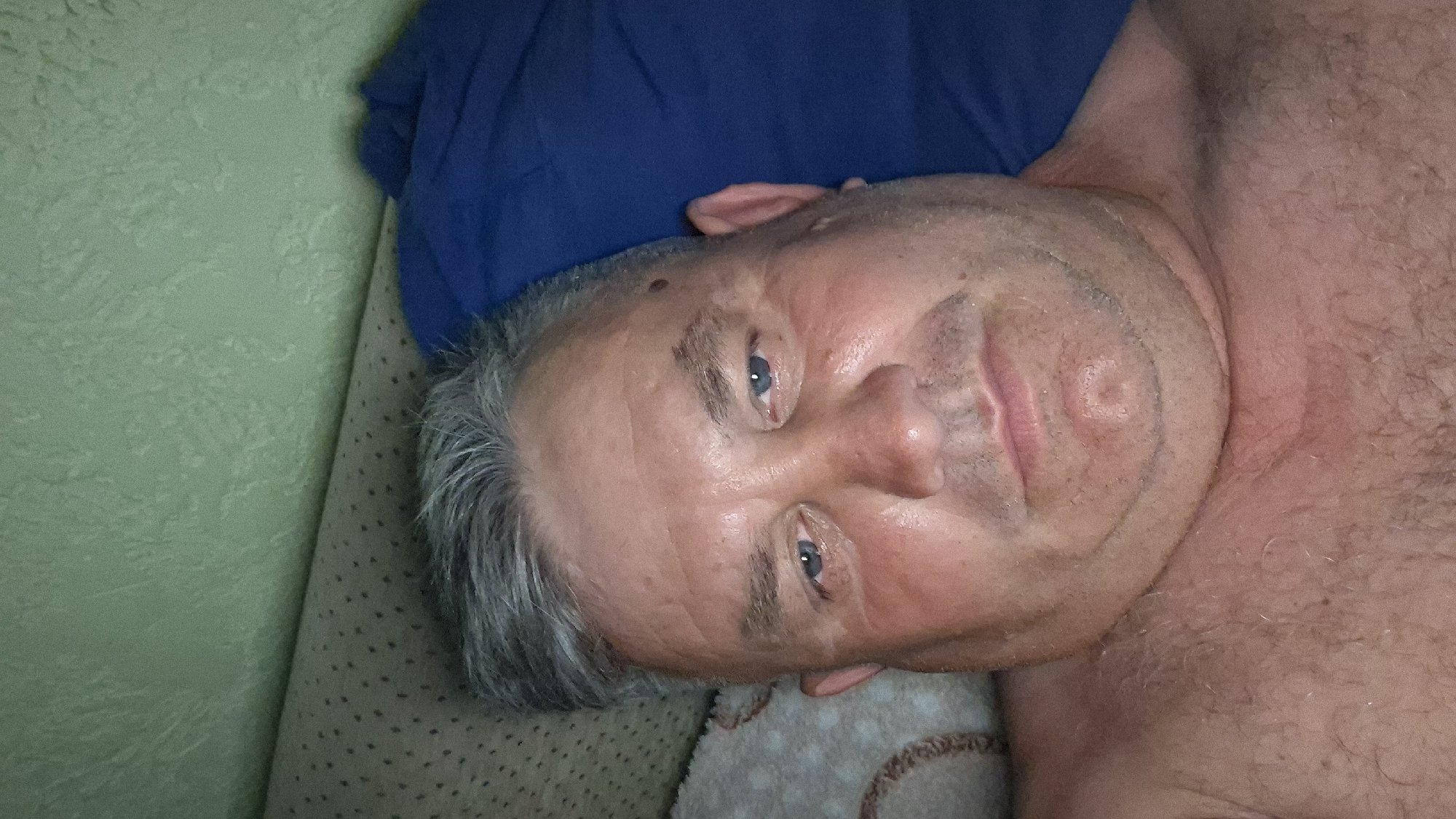 A man with grey hair and blue eyes lies on his back in bed. He is wearing a blue pillow and a green textured wall is visible behind him. The man appears to be in his late 50s or early 60s, with a round face and a double chin. He is looking directly at the camera with a neutral expression. The lighting is dim, suggesting it may be nighttime or a dimly lit room. The overall scene is intimate and personal, as if captured in a moment of relaxation or sleep.