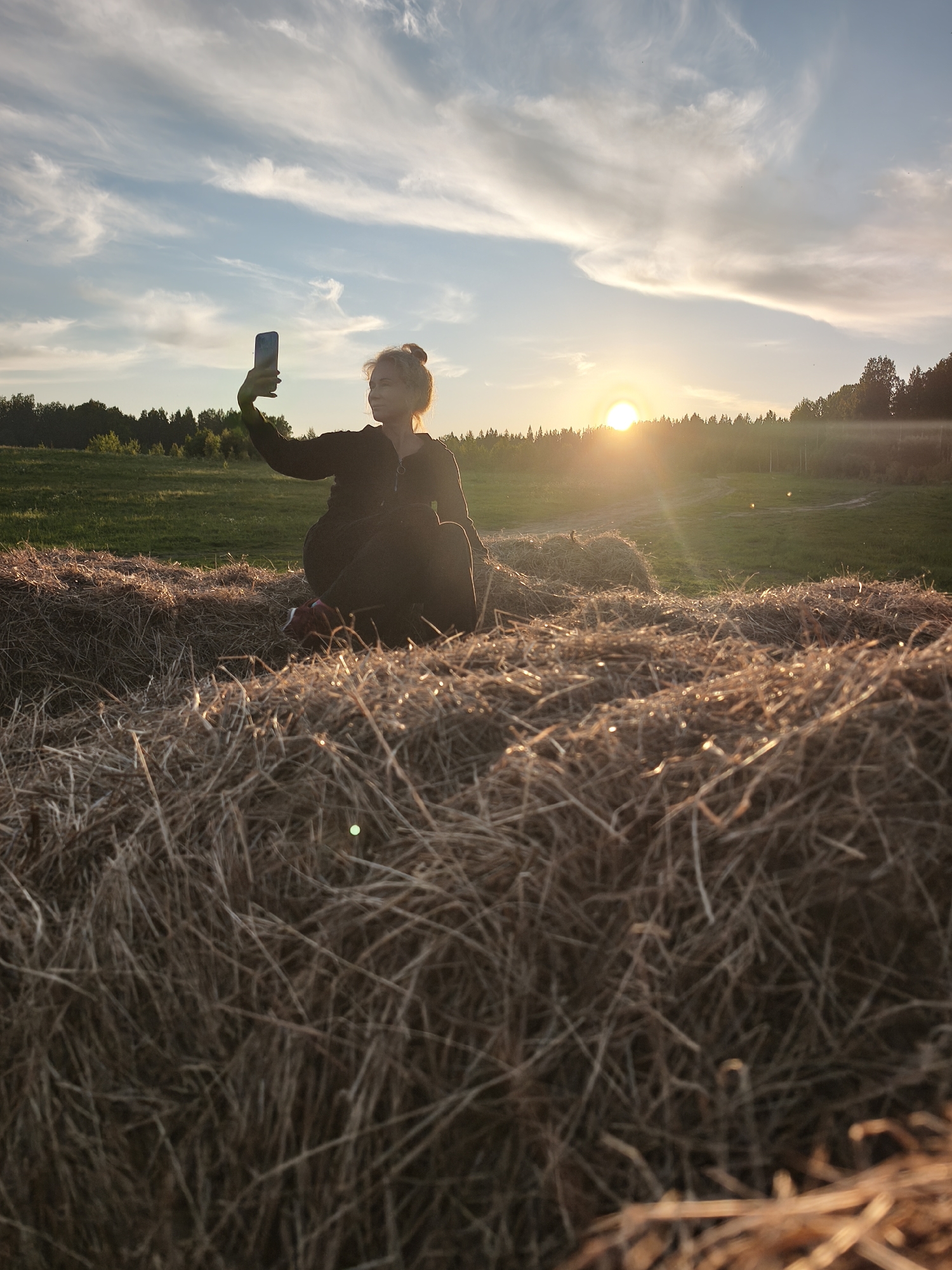 A wide outdoor shot captures a young woman sitting on a large hay bale in a sun-drenched field in Стеклянный, Russia, at what appears to be sunset or golden hour. The woman, positioned towards the left side of the frame, is holding a smartphone in her raised left hand, pointing it towards the distant landscape, suggesting she is taking a photo or a selfie. She is seen in profile, wearing dark clothing, possibly a hooded top, with her hair tied up. The warm, golden light of the setting sun illuminates her from behind, creating a partial silhouette and a soft glow around her form.
The sun itself is a bright, intense orb on the right side of the image, resting just above a distant treeline, and casts prominent lens flares into the scene. The sky is a blend of pale blue and soft orange, adorned with delicate, wispy white clouds that stretch across the upper portion of the frame. Below the treeline, a green field extends, indicating a rural or pastoral setting. The foreground is dominated by the intricate textures of dried hay bales, with the straw catching the warm light, adding depth and a rustic feel to the composition. The overall mood is peaceful and scenic, emphasizing the beauty of the natural landscape bathed in the late afternoon sun. No visible text is present in the image.
