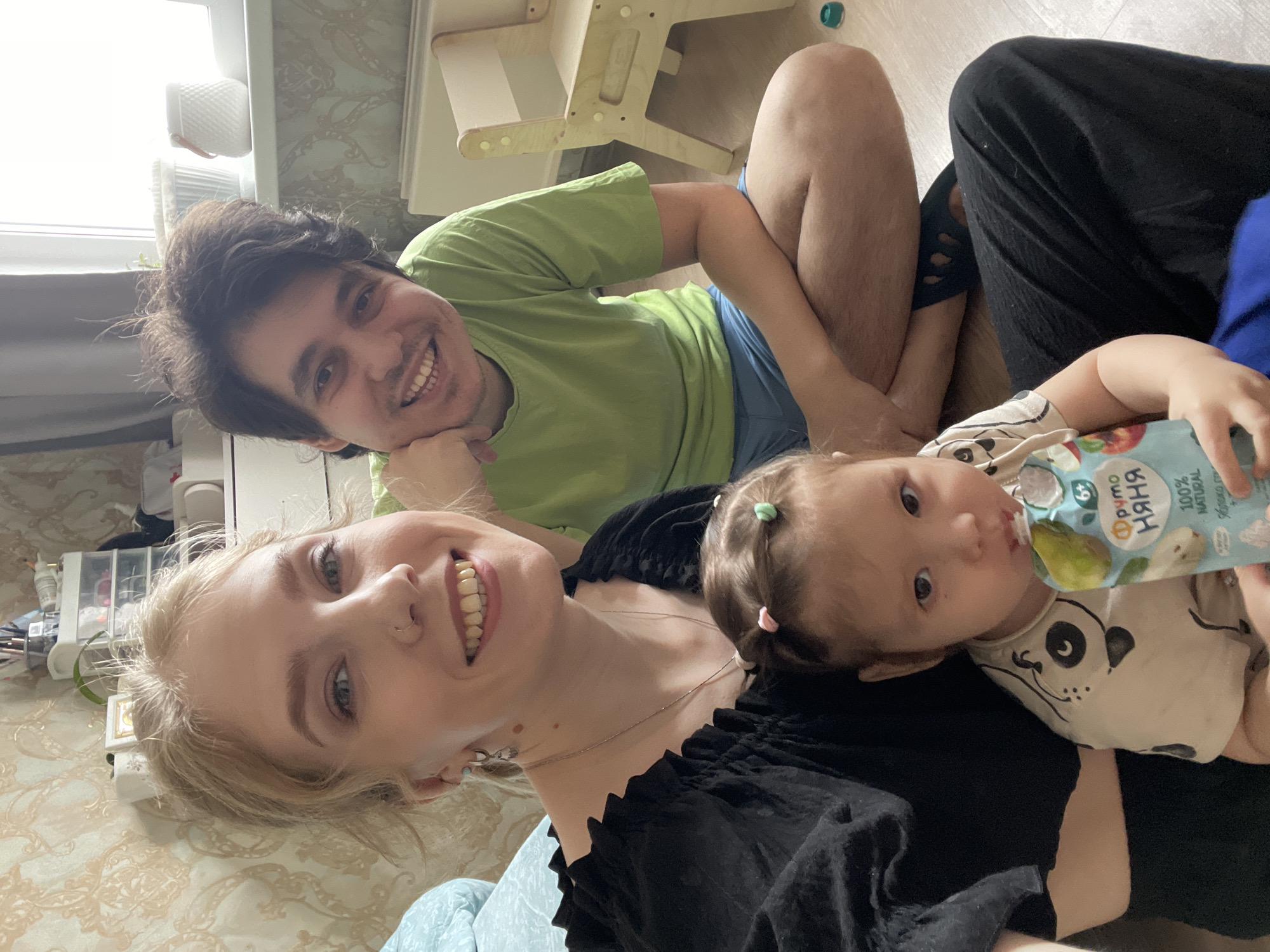 A joyful family of three, consisting of a man, a woman, and a young child, is captured in a bright indoor setting, likely a home in Izhevsk, Russia. All three subjects are smiling broadly at the camera, suggesting a happy, relaxed moment.
The man, appearing to be in his 20s or 30s with dark brown hair, wears a light green t-shirt and blue shorts. He is lying on his side, propping himself up on his elbow. Beside him, the woman, also appearing to be in her 20s or 30s with blonde hair and a nose piercing, wears a black top and smiles directly at the viewer. A young child, likely a baby or toddler with light brown hair styled with small pink and green hair ties, rests on the woman's chest. The child is wearing a white t-shirt with a cartoon panda face and is actively drinking from a blue, white, and green food pouch.
The food pouch clearly displays Russian text, including "Фруто няня" (Fruto Nyanya), a common baby food brand, "100% NATURAL," "6+" (indicating suitability for babies over 6 months), and "БЕЗ ГМО" (GMO-free), alongside an illustration of a pear.
The setting features a bright window in the background, indicating daytime. Light-colored wooden flooring and a patterned rug or bedspread are visible. A small wooden children's chair or step stool is partially seen near the man, and a container with various small items sits near the woman. The overall atmosphere is warm and intimate, capturing a candid family moment.