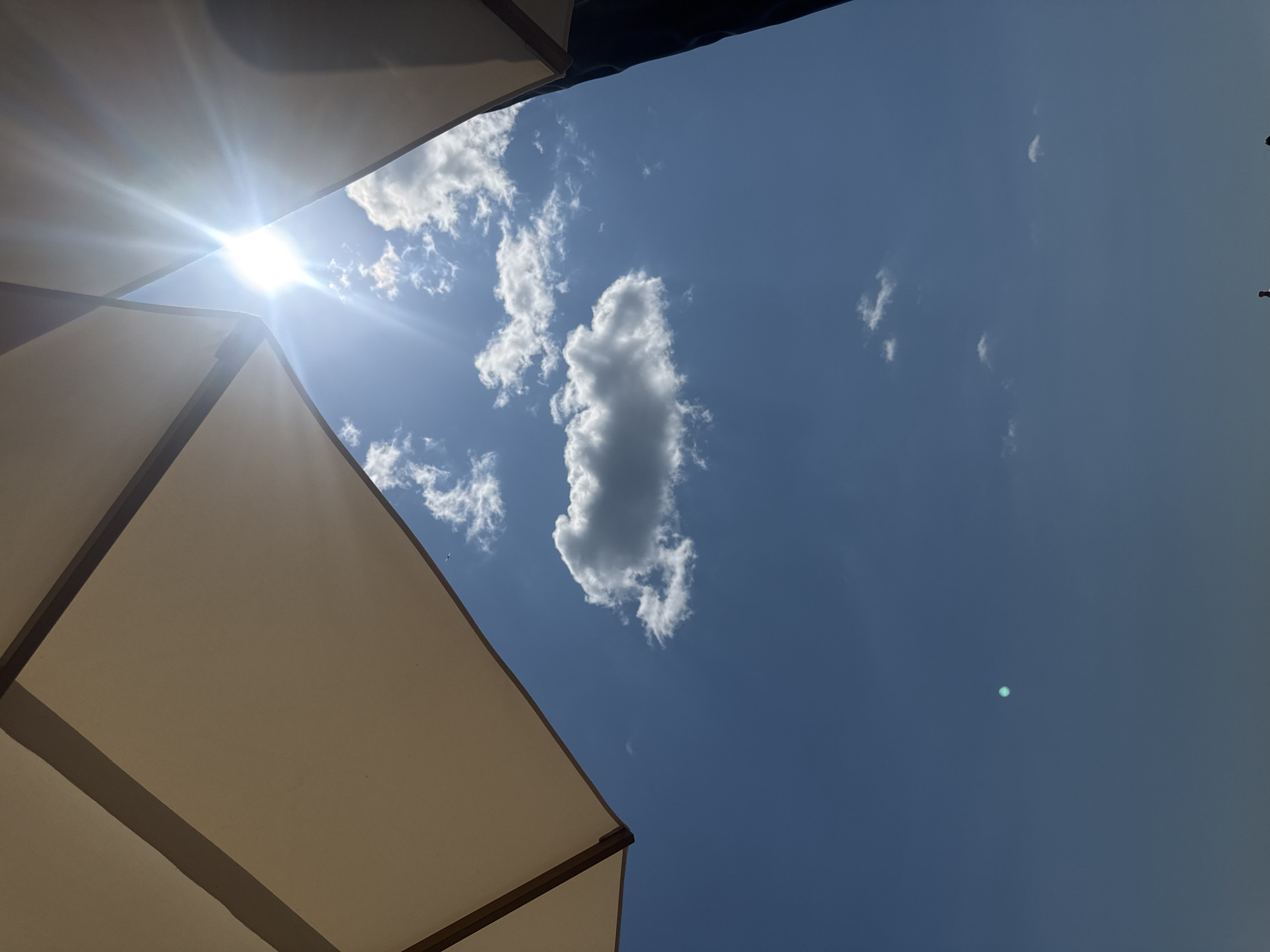 The image shows a view looking up at a clear blue sky with a few scattered clouds. The sun is shining brightly, creating lens flare. The edges of two large, light-colored umbrellas are visible at the top of the frame, suggesting an outdoor seating area. The bottom of the image is dark and indistinct, possibly showing people or structures below. A small, bright turquoise light is visible in the sky, which could be a lens artifact or a distant object. The scene is not specific to Odesa, Ukraine, as there are no identifying landmarks or contextual clues.