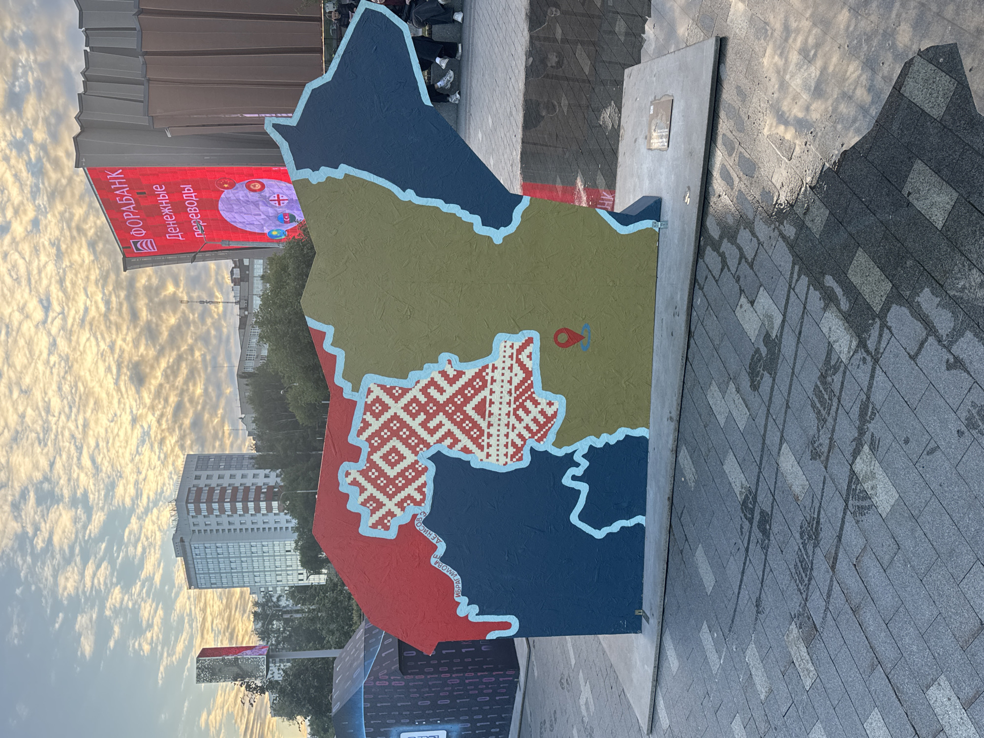 The image captures an outdoor scene in Perm, Russia, featuring a large, colorful map-shaped art installation. The map is divided into different colored regions: red, blue, and tan. A distinct section of the map is adorned with a traditional Belarusian embroidery pattern in red and white. A red location pin marker is visible on the tan portion of the map.
In the background, a large red banner displays text in Russian. The text reads "ФОРАБАНК" (FORABANK) and "Денежные переводы" (Money transfers). Several circular emblems, possibly flags, are also visible on the banner. To the left of the banner, a modern building with many windows and a striped facade can be seen against a sky filled with dramatic, golden-hour clouds. The time of day appears to be late afternoon or early evening, indicated by the warm sunlight and the soft shadows.
The art installation is placed on a concrete base, which in turn rests on a paved area. The paving stones are laid in a diagonal pattern, and some appear wet, reflecting the sky and surrounding structures. There are faint tire tracks visible on the wet pavement. A few people are standing in the background, near a dark, textured wall. The overall atmosphere suggests a public space, possibly a plaza or an exhibition area, where art and advertising converge.