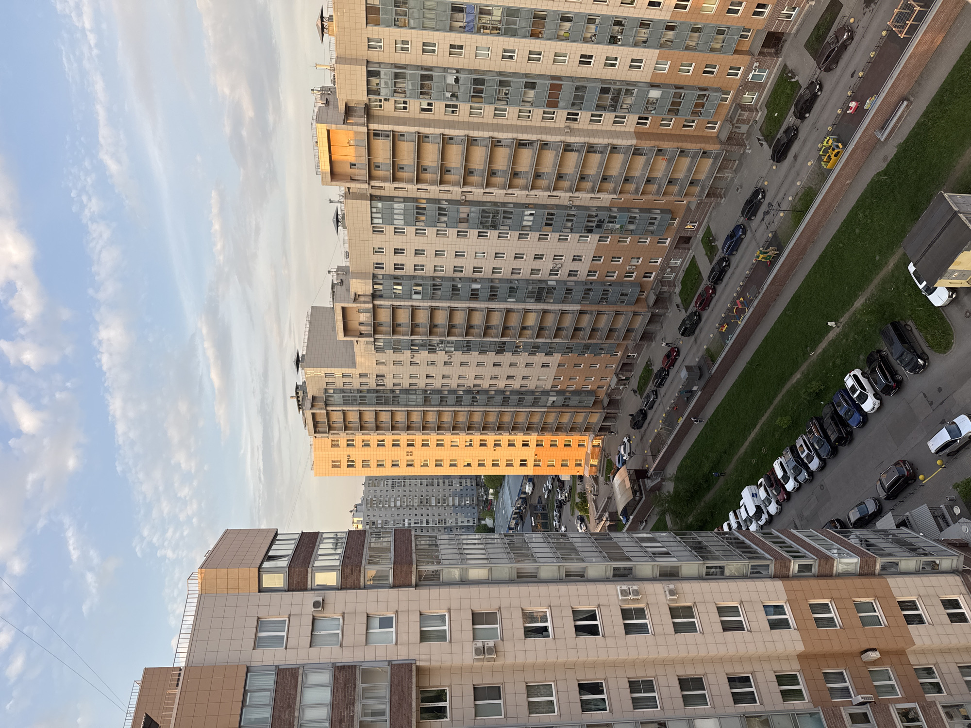 The image captures a high-angle view in Sinyavino, Russia, showcasing a cluster of residential buildings against a partly cloudy sky. The buildings exhibit modern architectural designs with varied heights and facades composed of light beige and brown tiles. Some have glass-paneled balconies. A road runs parallel to the buildings, lined with parked cars of various colors, primarily black and white. Some greenery, likely grass verges, is visible alongside the road. The lighting suggests late afternoon or early morning, with some buildings reflecting sunlight. Several pedestrians and two small yellow vehicles are walking along the road next to the buildings. In the distance, more high-rise buildings are visible.