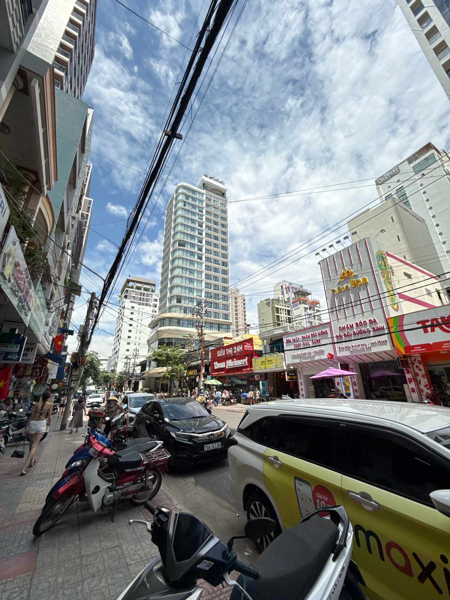 The image shows a street scene in Nha Trang, Vietnam. On the right side of the street, there is a white car with yellow and red branding that says "maxi." Next to it, there are several motorcycles parked on the sidewalk. Further down the street, there are shops and tall buildings. The sky is blue with white clouds. The street is busy with traffic, including cars and motorcycles. People are walking on the sidewalk and some are riding motorcycles. One person is wearing a conical hat, which is a traditional Vietnamese hat. The overall impression is of a bustling city street on a sunny day.