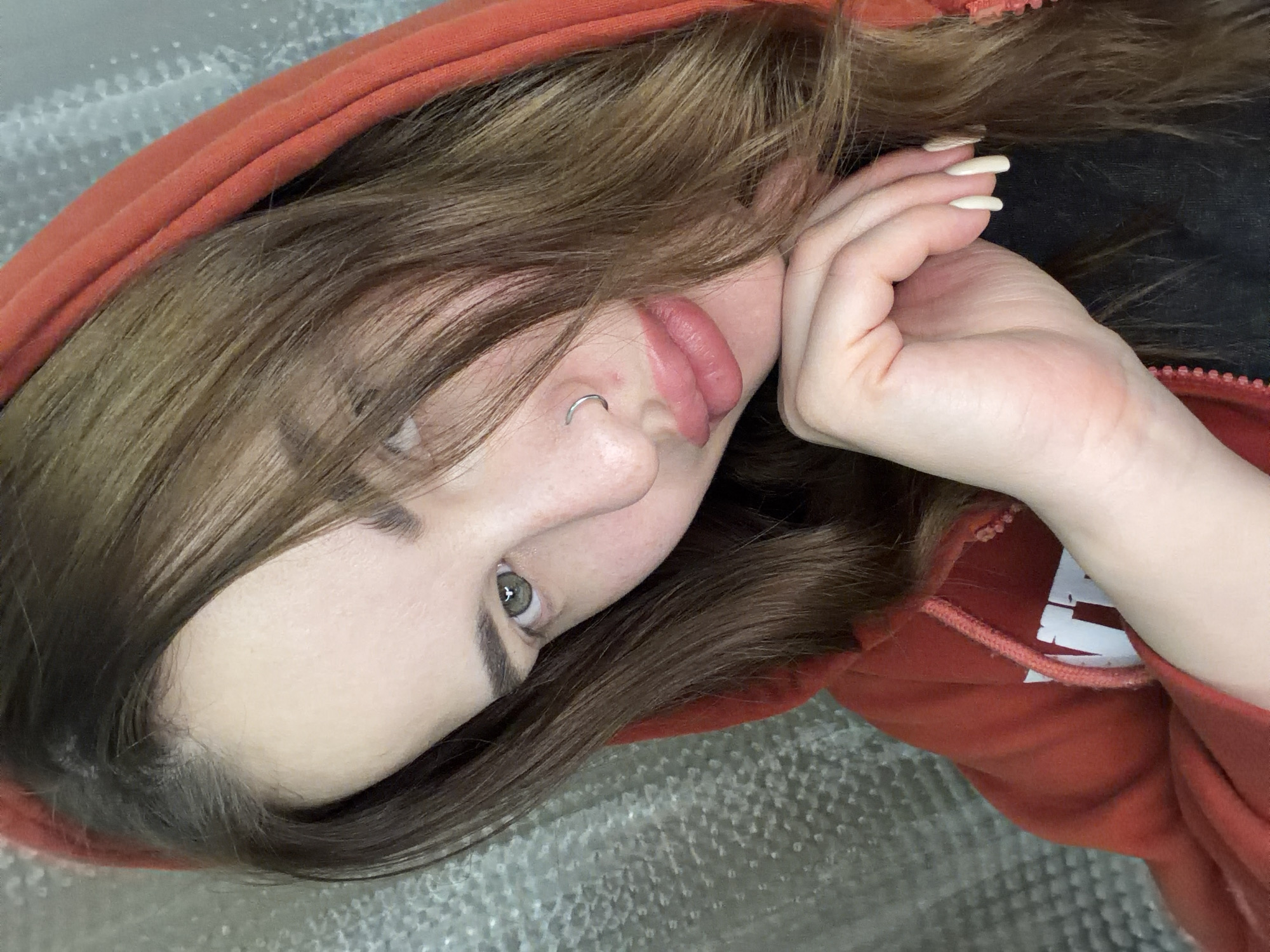 A young woman is reclining, wearing an orange hooded sweatshirt. She has brown hair, light skin, and a nose ring. Her manicured hand is near her face. The background appears to be a silver-colored textured surface. No information is present to indicate the image was taken in Krasnodar, Russia.
