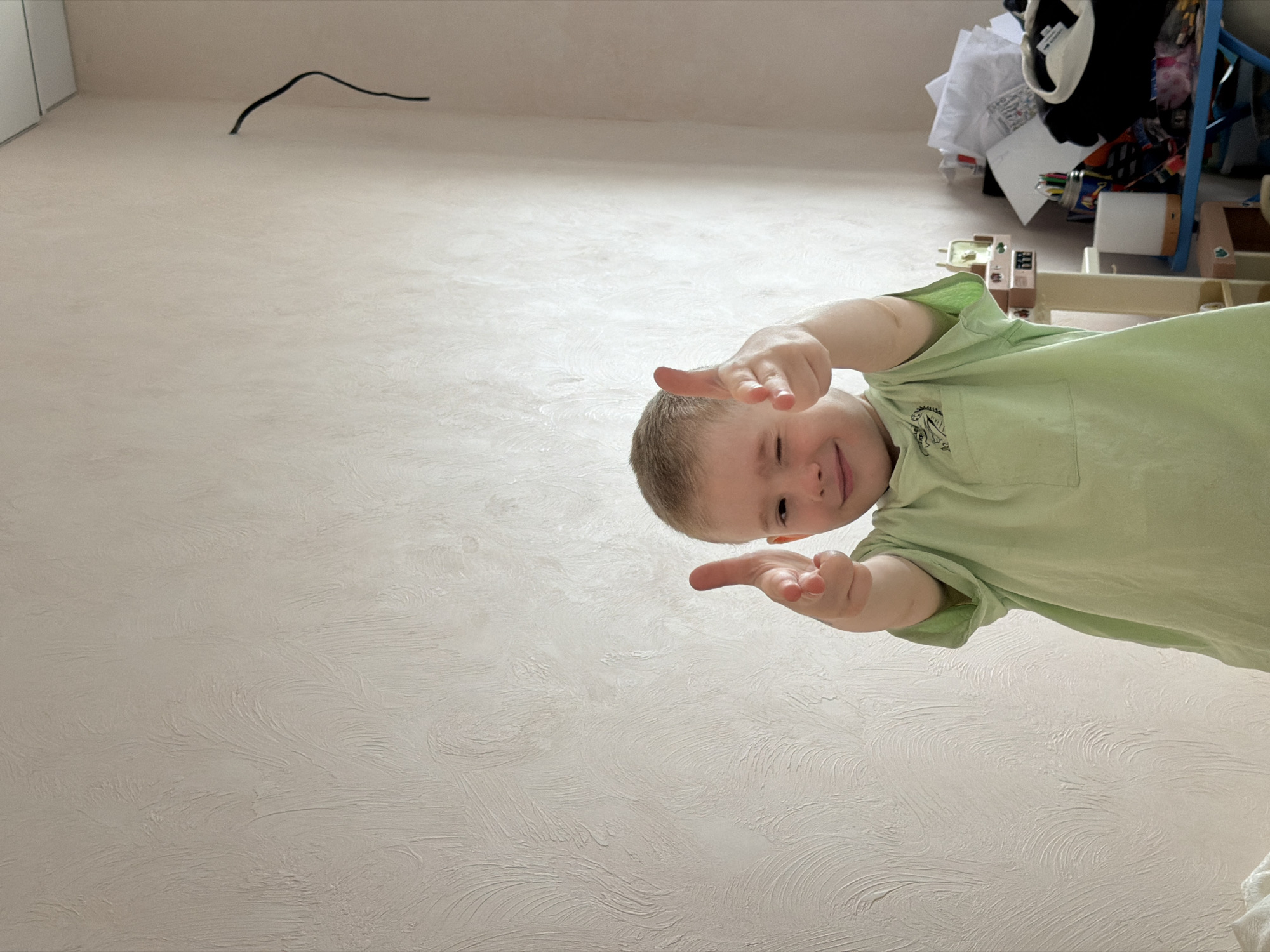 A boy with short, light-brown hair stands facing the camera, wearing a light-green t-shirt with a cartoon dinosaur on the pocket. He is smiling and pointing his index fingers and thumbs towards the camera. The background is a light-beige wall with textured patterns, along with a dark cord hanging from the wall. To the upper right is a cluttered shelf with clothing, paper, and objects on and around it. The photo appears to have been taken from a low angle. The city is Rossiyskiy, Russia.