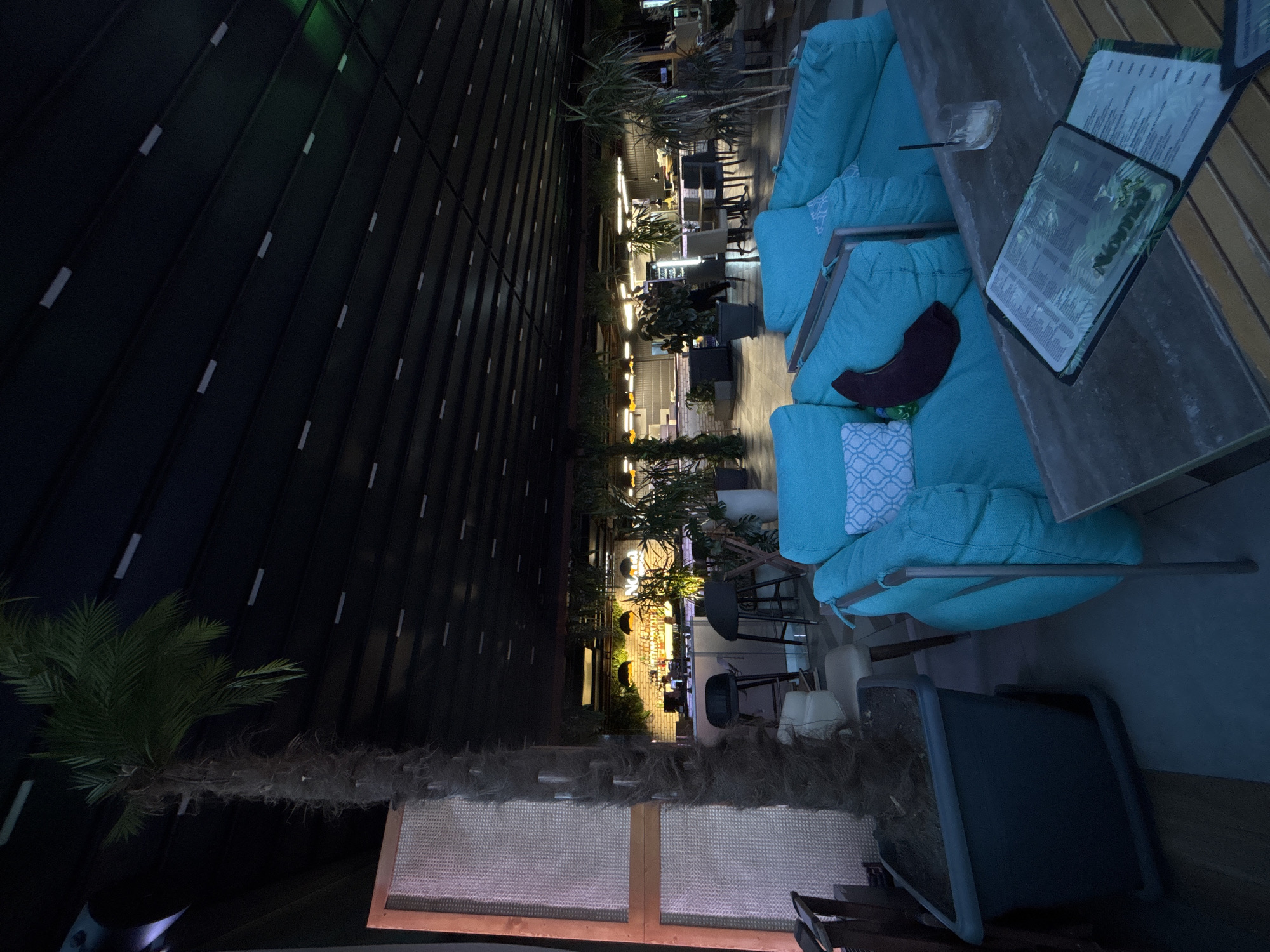 The image depicts an outdoor seating area at a restaurant or bar in Yerevan, Armenia, during nighttime. In the foreground, a large grey planter contains a tall palm tree. To the right, a section of turquoise outdoor seating furniture, possibly beanbag chairs, is visible. A wooden table is situated in front of the seating, displaying a menu with a tropical leaf design and a glass with a straw and ice.
In the background, a bar area is illuminated with warm lighting. The bar is lined with bottles and appears to be staffed, with at least one person visible behind the counter. Various potted plants are strategically placed throughout the establishment, adding to the ambiance. The ceiling above is a dark, slatted structure with recessed lighting fixtures. The overall impression is that of a stylish, contemporary outdoor dining and drinking venue.