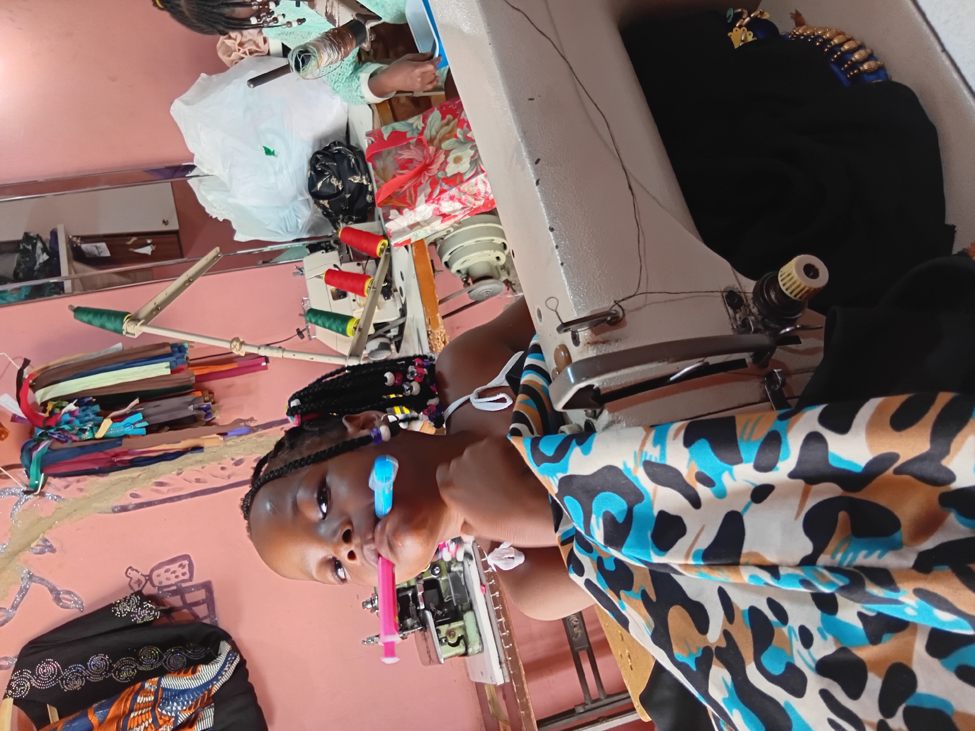 A young Black girl with braided hair is sitting at a sewing machine, with a pink and blue object in her mouth. She is wearing a white tank top with a patterned skirt or fabric draped in front of her. The background shows a wall painted pink, with various items related to sewing such as spools of thread, fabric, a sewing machine, and a mirror. A large white bag is also visible, along with a red floral patterned bag. The overall scene suggests a workspace, possibly a tailor's shop or a home sewing area.