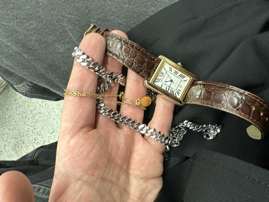 The image displays a close-up, downward-angled view inside a London Underground train carriage, in London, United Kingdom. A person's bare left hand, resting on dark-colored trousers, holds multiple items of jewelry. The primary object is a rectangular, gold-toned Cartier Tank watch, featuring a white dial with black Roman numerals and a date display showing "25." The watch face also indicates "AUTOMATIC." It is attached to a worn, brown, alligator-textured leather strap. Additionally, the hand holds a silver-colored, curb-link chain and a finer gold-colored chain from which a small, round, textured, orange-brown pendant is suspended. The background shows the characteristic speckled grey and blue floor pattern of a London Underground train.