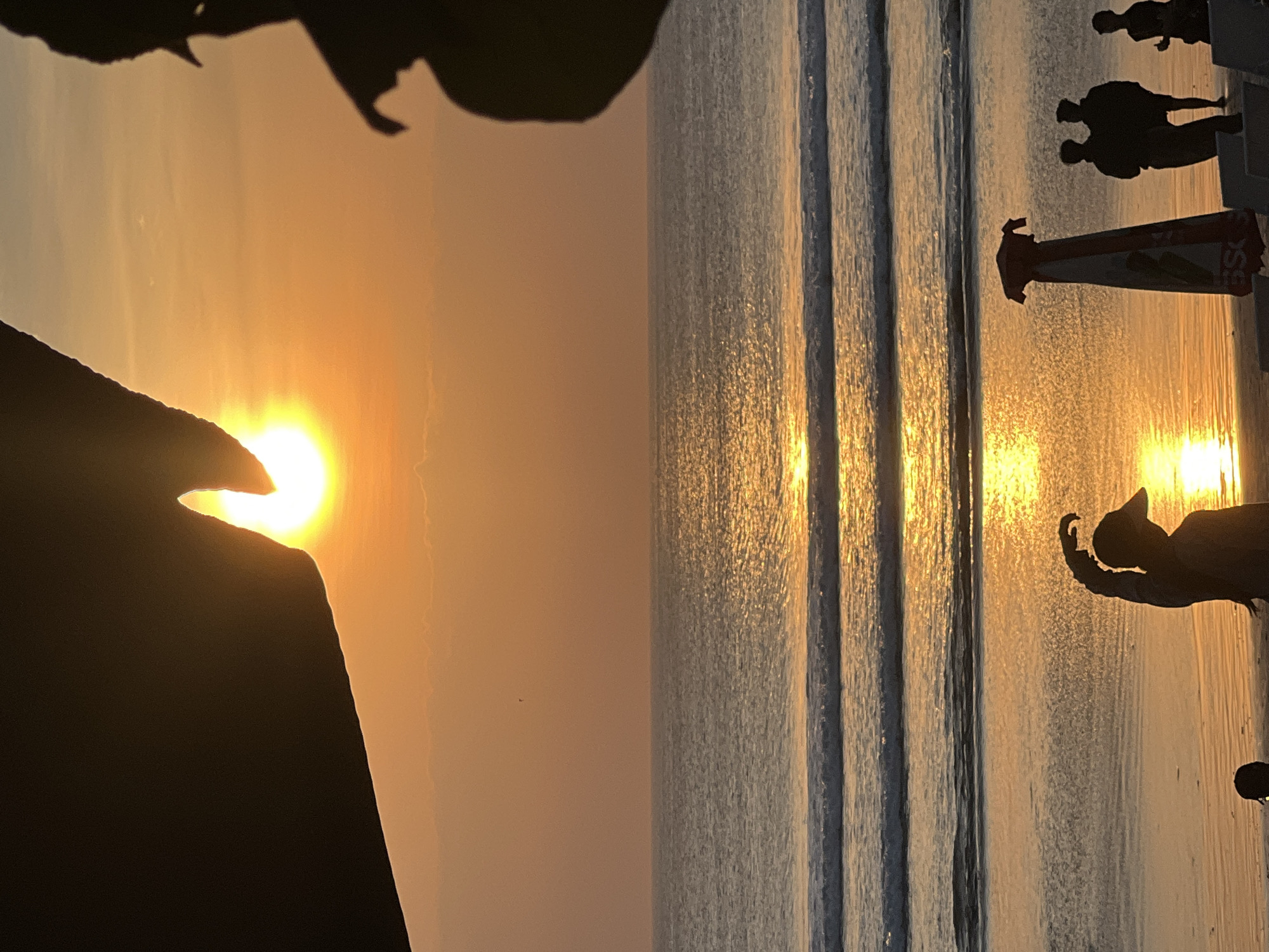 The image depicts a sunset over the ocean at Kuta Beach, Bali, Indonesia. The sun, a bright orb of orange, is partially obscured by dark silhouetted structures in the foreground and mid-ground. The sky is a gradient of orange and yellow, with a few wispy clouds. The ocean is calm, with gentle waves rolling onto the sandy shore. The water reflects the warm colors of the sunset, creating a shimmering path of light.
On the beach, several people are visible as dark silhouettes. In the foreground, a person wearing a baseball cap is facing the ocean, with their arm raised as if waving or gesturing. To the right, a tall, umbrella-like object stands on the sand. Further back, two people stand close together, and another individual is visible to their right. The beach itself appears to be sandy, with some lounge chairs in the lower right corner of the frame. The overall scene is tranquil and picturesque, characteristic of a beach sunset in Bali.