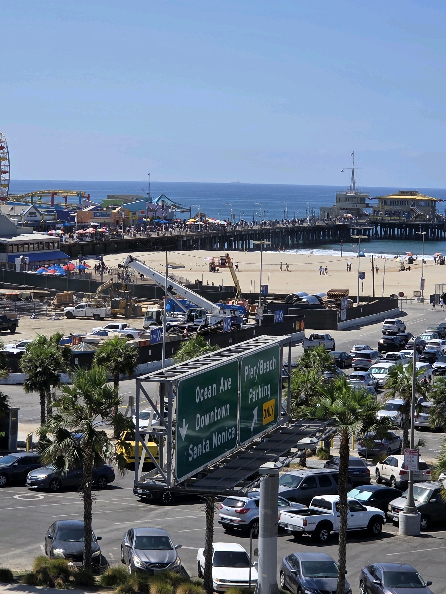 This is a daytime, high-angle, full shot of the Santa Monica Pier and beach area in Santa Monica, California, United States of America. The scene is bustling with activity. In the foreground, a parking lot is filled with various cars, including sedans, SUVs, and pickup trucks. Palm trees are interspersed throughout the parking lot. A large green directional sign for "Ocean Ave Downtown Pier/Beach Parking" is prominently displayed. Further back, construction is underway with cranes and heavy machinery visible. Beyond the construction, the Santa Monica Pier extends into the ocean, crowded with people and featuring amusement park rides like a Ferris wheel and a roller coaster. The beach is populated with sunbathers and individuals walking along the shore. The ocean stretches to the horizon under a clear blue sky.