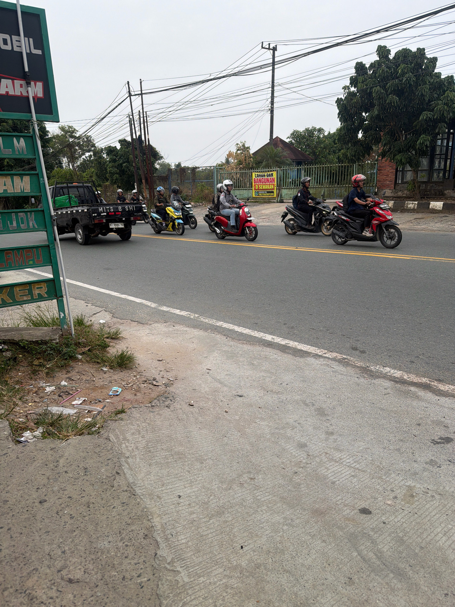 In Balikpapan, Indonesia, a group of motorcyclists ride along a paved road. The scene is captured on an overcast day, with a pale sky overhead. On the left side of the road, a green signpost stands, displaying white text that reads "DOBEL", "ARM", "LM", "AM", "JOK", "AMPU", and "KER". To the right of the signpost, a black pickup truck with several passengers is visible. Further down the road, a procession of five motorcycles, each with one or two riders wearing helmets, proceeds in the same direction. The riders are dressed in casual attire. The road is lined with trees and vegetation on the right side, with a metal fence and buildings in the background. A yellow banner with Indonesian text is also visible on the right side of the road. The foreground shows a concrete sidewalk and a patch of dirt with scattered debris. The asphalt road features double yellow lines, indicating traffic lanes.