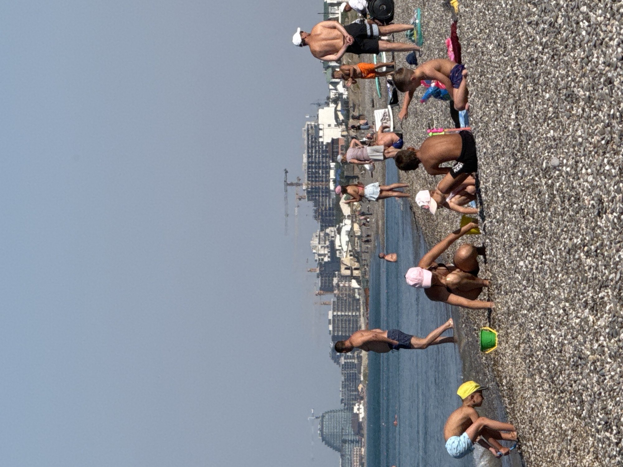 The image depicts a sunny day on a pebble beach in Novofedorovka, Ukraine. A crowd of people are enjoying the sea and the shore. In the foreground, two young children are playing in the shallow water with a green bucket and toys. A woman in a bikini and a pink cap is also squatting on the beach near them. Further back, several adults and children are scattered across the beach, some standing, some sitting, and some wading in the sea. The background reveals a cityscape with modern buildings under construction, including a distinctive curved glass building, and cranes indicating ongoing development. The sea is calm and clear, with a few people swimming in the distance. The sky is a bright, cloudless blue.